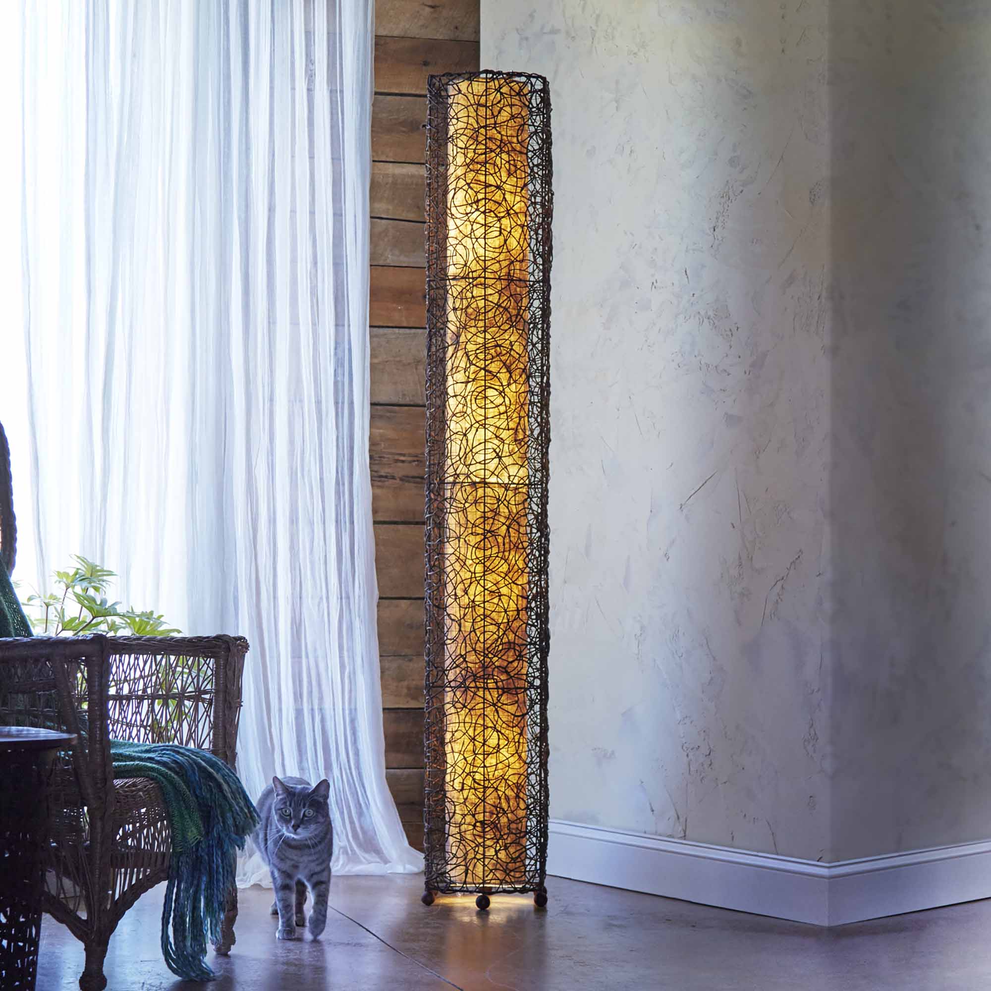 In a softly lit reading nook, crafted by the artisans of Eangee Home Design, the Nito Giant Floor Lamp in natural stands gracefully, featuring recycled paper shade, eco friendly, sustainably sourced, recycled materials, adding warmth and character to the space.