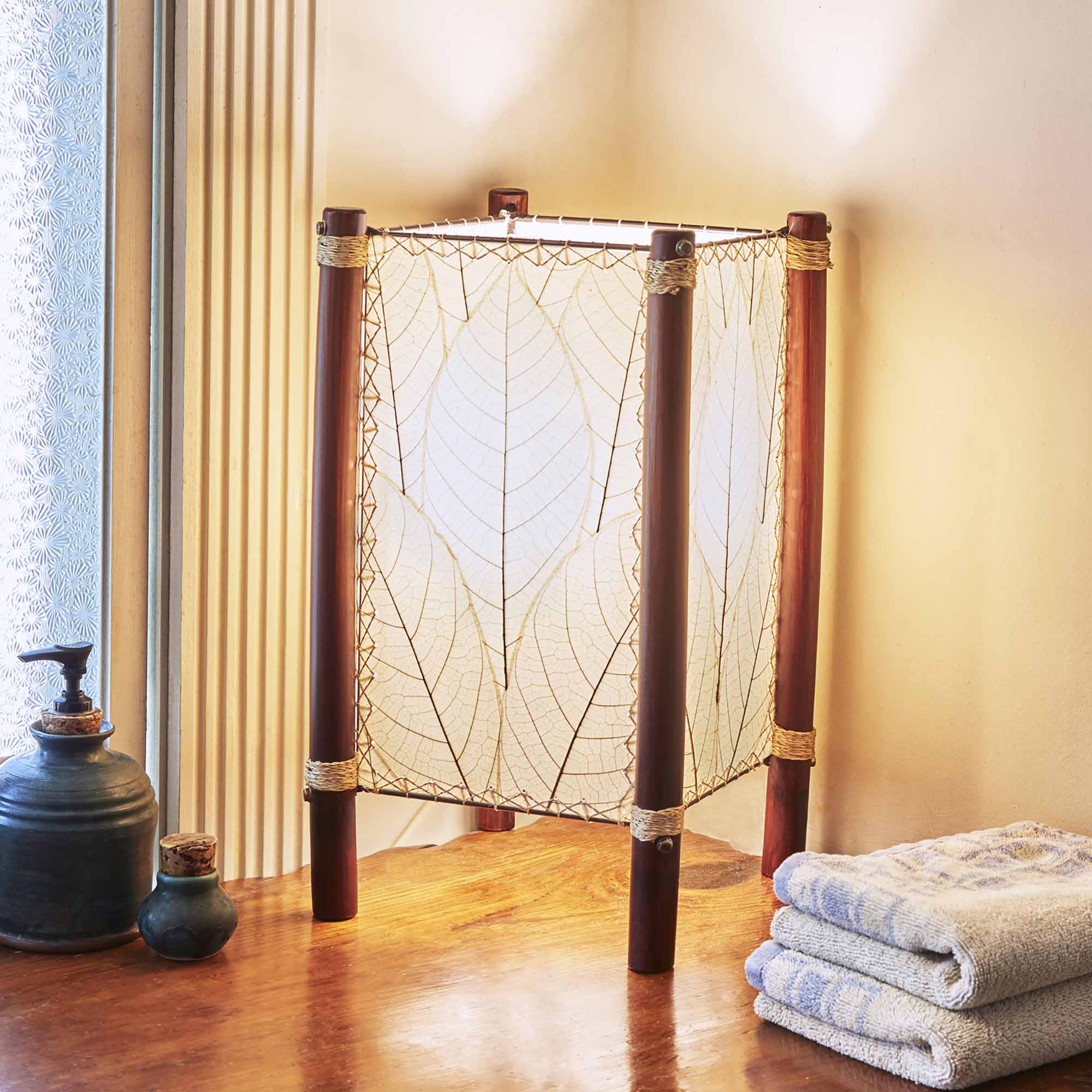 In a minimalist loft with natural textures, crafted by the artisans of Eangee Home Design, the Fortune Table Lamp in natural stands gracefully, featuring fossilized cocoa leaf shade, abaca rope accents, rattan leg supports, adding warmth and character to the space.