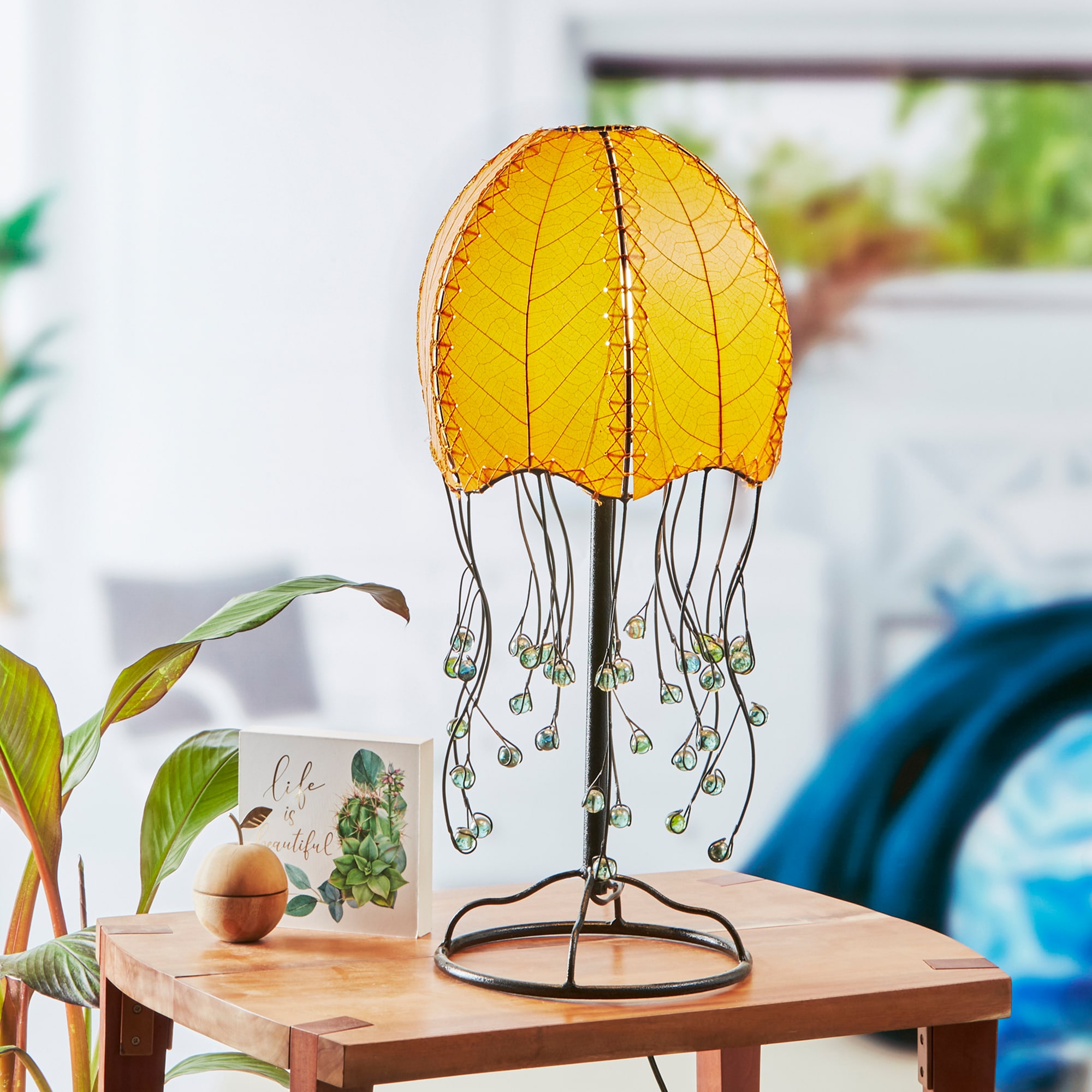 In a quiet bedside corner, crafted by the artisans of Eangee Home Design, the Jellyfish Table Lamp in orange stands gracefully, featuring fossilized cocoa leaf shade, abaca rope accents, wrought iron frame, handcrafted, adding warmth and character to the space.