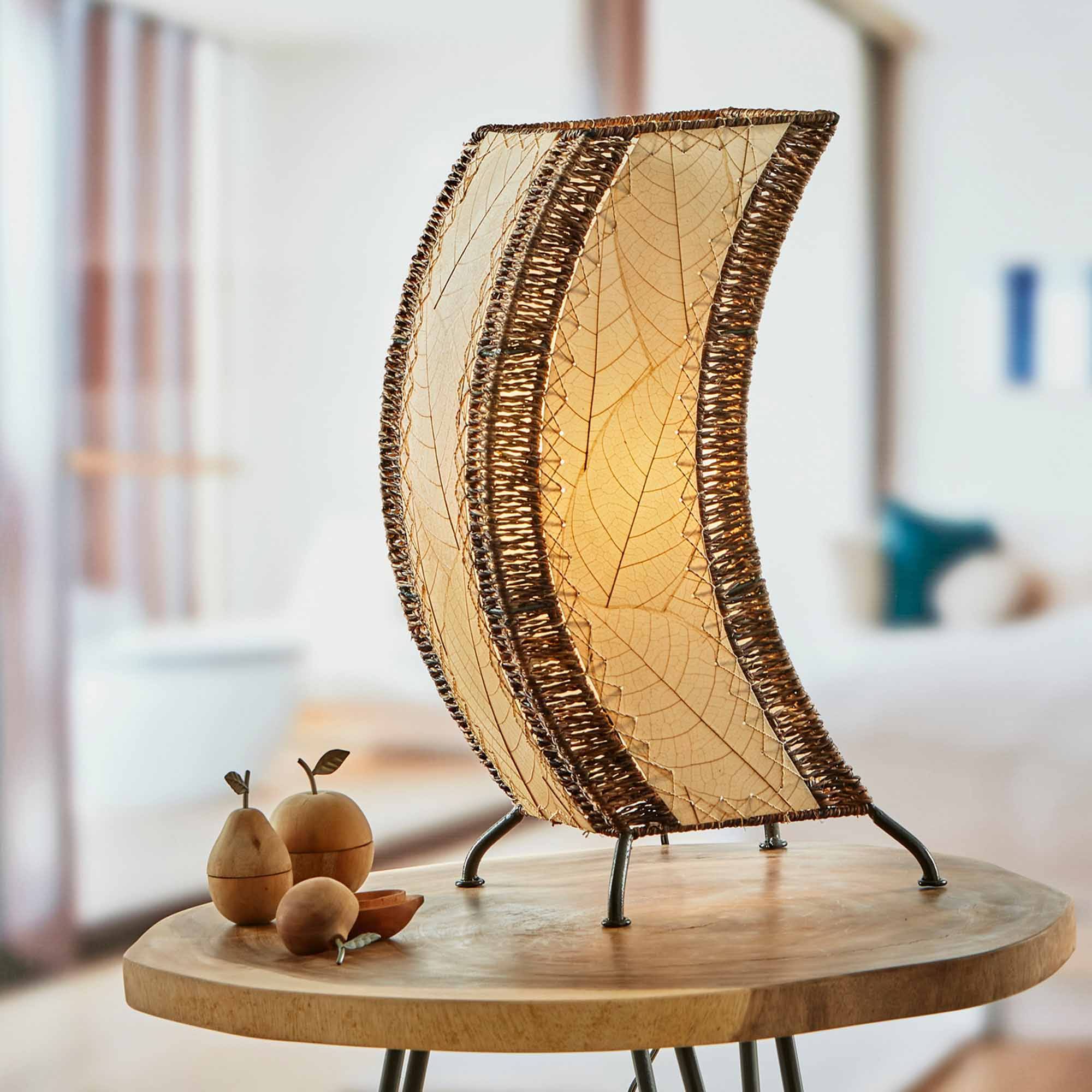 In a softly lit reading nook, crafted by the artisans of Eangee Home Design, the C Shape Table Lamp in natural stands gracefully, featuring fossilized cocoa leaf shade, abaca rope accents, fair trade, handcrafted, adding warmth and character to the space.