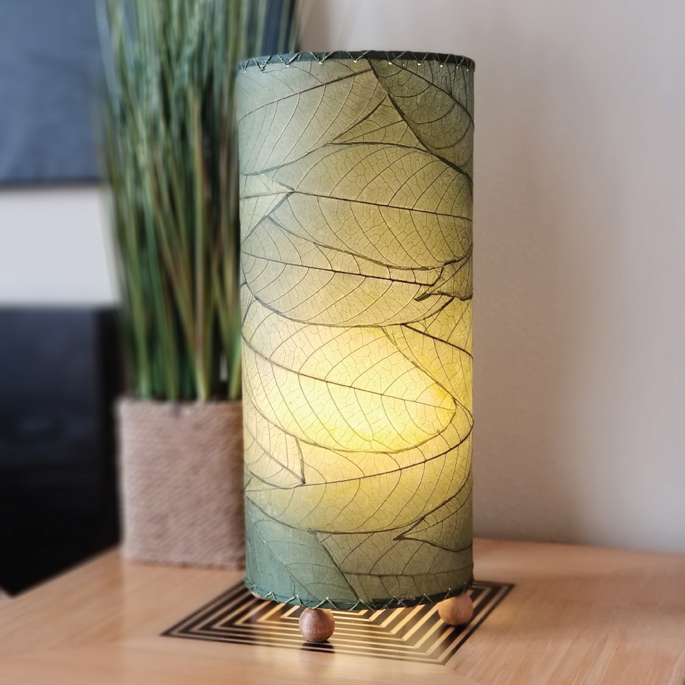 Crafted by the artisans of Eangee Home Design, the Cocoa Leaf Cylinder Table Lamp in green against a clean backdrop, featuring fossilized cocoa leaf shade, wrought iron frame, handcrafted.