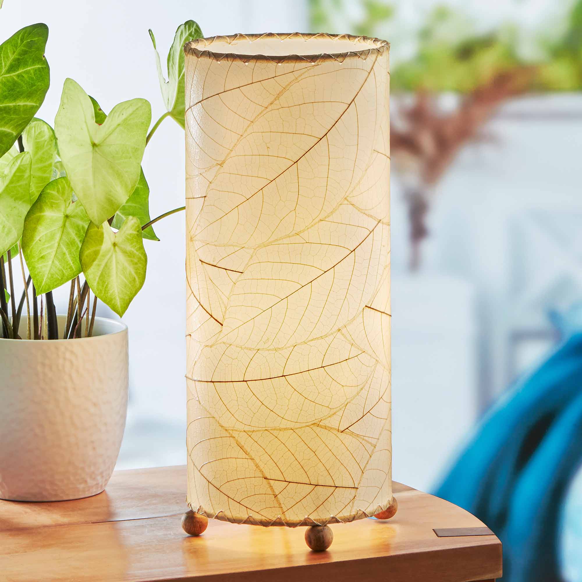 In a softly lit reading nook, crafted by the artisans of Eangee Home Design, the Cocoa Leaf Cylinder Table Lamp in natural stands gracefully, featuring fossilized cocoa leaf shade, wrought iron frame, handcrafted, adding warmth and character to the space.