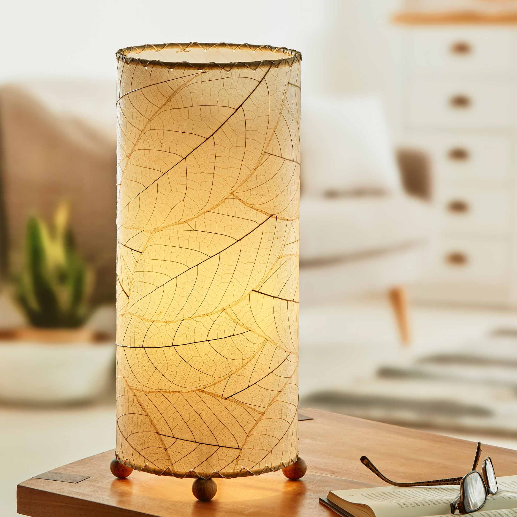 In a sun drenched entryway, crafted by the artisans of Eangee Home Design, the Cocoa Leaf Cylinder Table Lamp in natural stands gracefully, featuring fossilized cocoa leaf shade, wrought iron frame, handcrafted, adding warmth and character to the space.