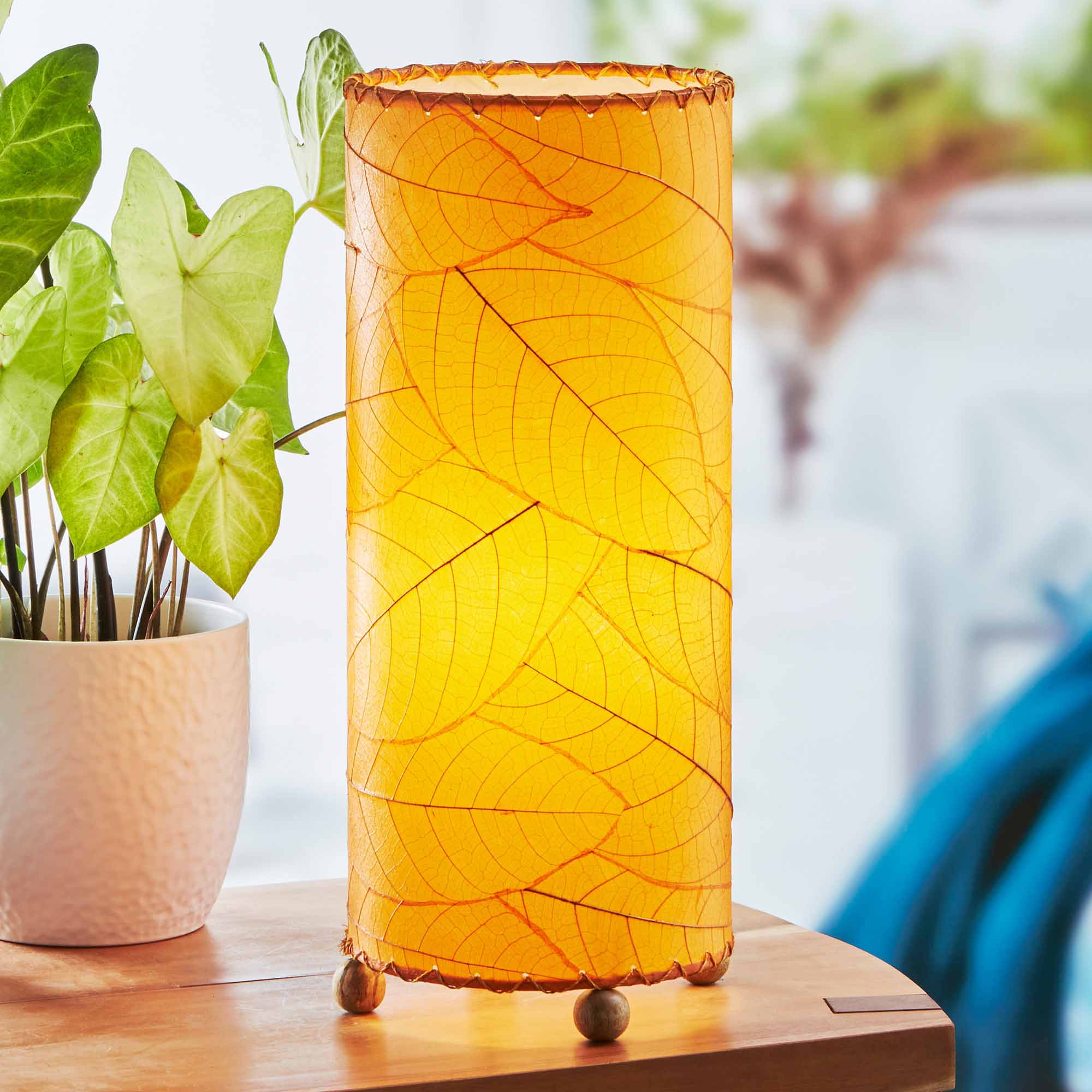 In a quiet bedside corner, crafted by the artisans of Eangee Home Design, the Cocoa Leaf Cylinder Table Lamp in orange stands gracefully, featuring fossilized cocoa leaf shade, wrought iron frame, handcrafted, adding warmth and character to the space.