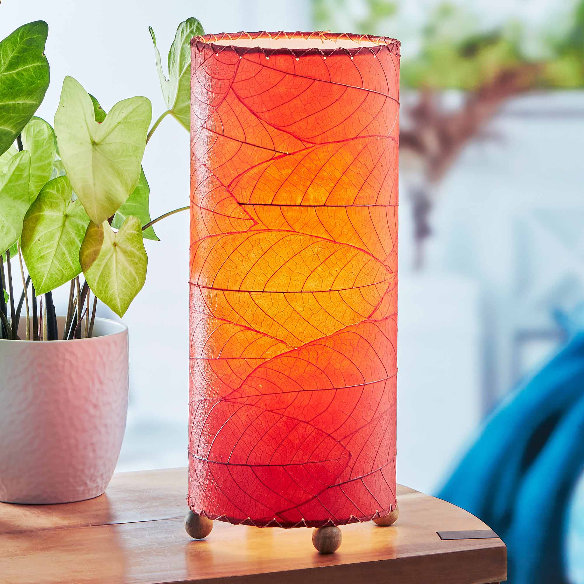 In a modern interior with earth tones, crafted by the artisans of Eangee Home Design, the Cocoa Leaf Cylinder Table Lamp in red stands gracefully, featuring fossilized cocoa leaf shade, wrought iron frame, handcrafted, adding warmth and character to the space.