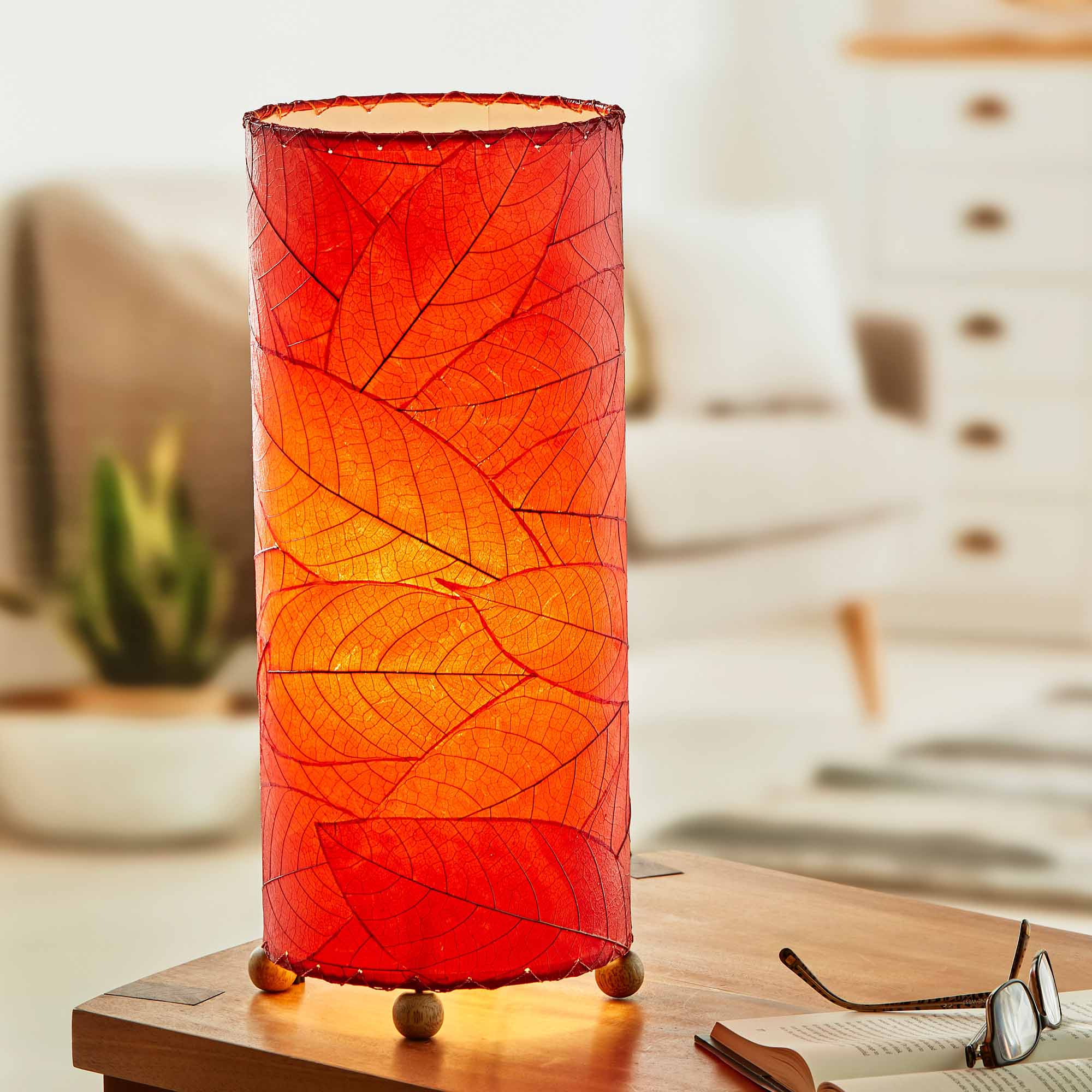 In a modern interior with earth tones, crafted by the artisans of Eangee Home Design, the Cocoa Leaf Cylinder Table Lamp in red stands gracefully, featuring fossilized cocoa leaf shade, wrought iron frame, handcrafted, adding warmth and character to the space.