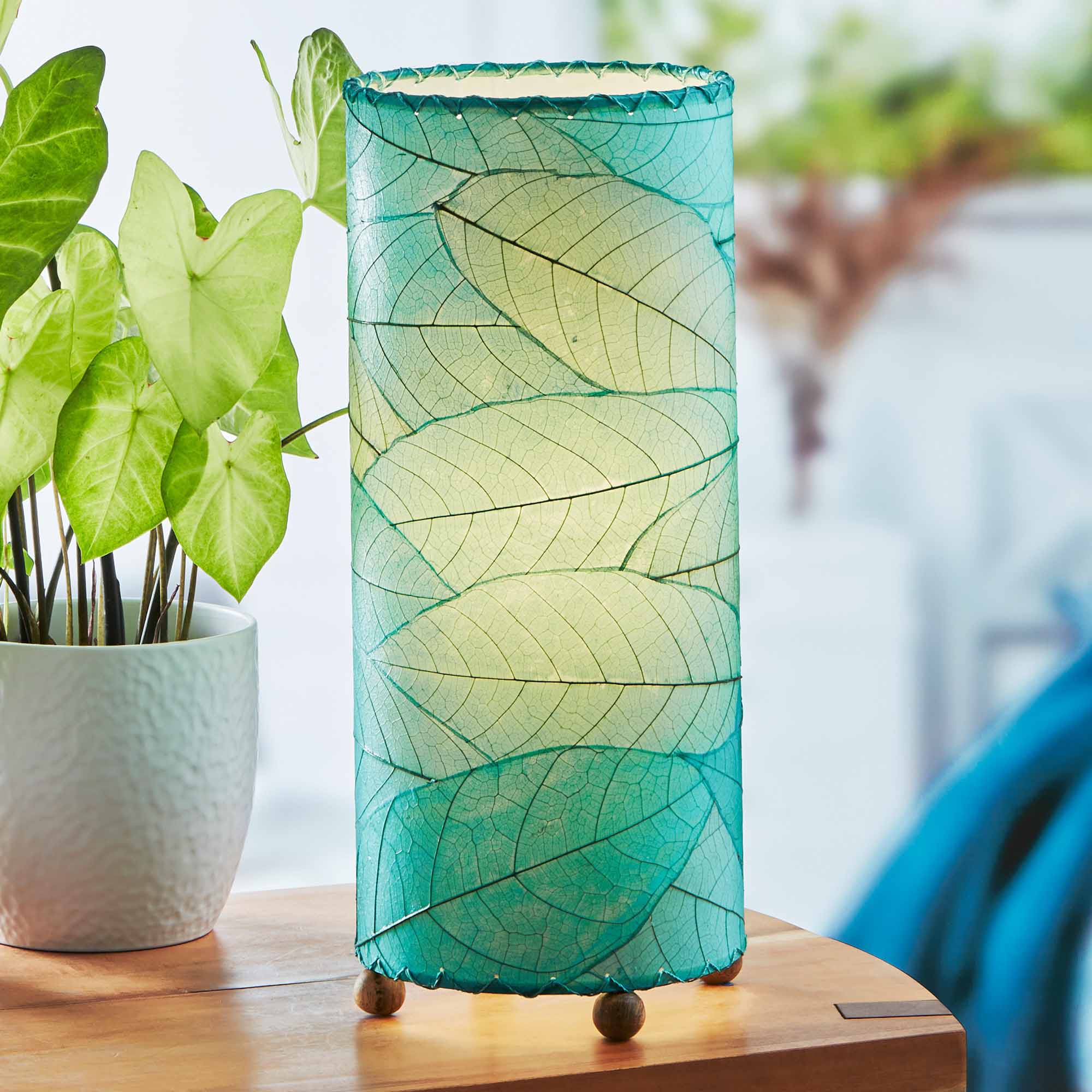 In a softly lit reading nook, crafted by the artisans of Eangee Home Design, the Cocoa Leaf Cylinder Table Lamp in sea blue stands gracefully, featuring fossilized cocoa leaf shade, wrought iron frame, handcrafted, adding warmth and character to the space.