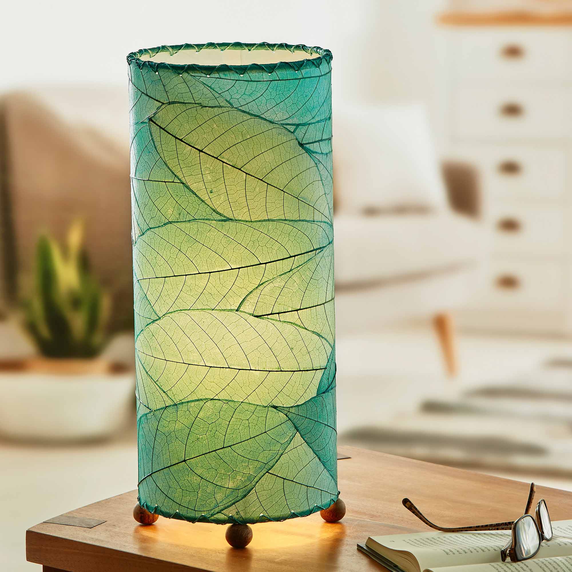 In a modern interior with earth tones, crafted by the artisans of Eangee Home Design, the Cocoa Leaf Cylinder Table Lamp in sea blue stands gracefully, featuring fossilized cocoa leaf shade, wrought iron frame, handcrafted, adding warmth and character to the space.