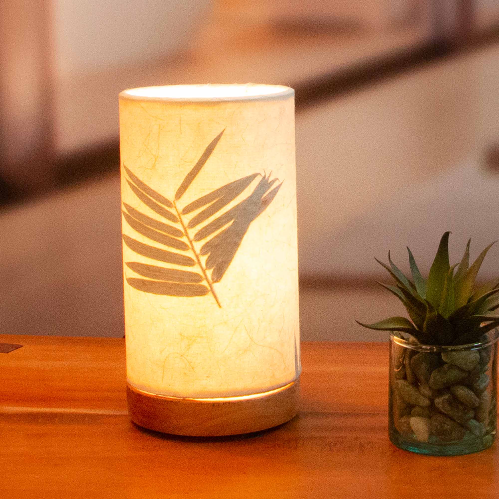 In a modern interior with earth tones, crafted by the artisans of Eangee Home Design, the Mini Paper Cylinder Table Lamp Bamboo in natural stands gracefully, featuring reclaimed mango wood base, recycled paper shade, sustainably sourced, recycled materials, adding warmth and character to the space.
