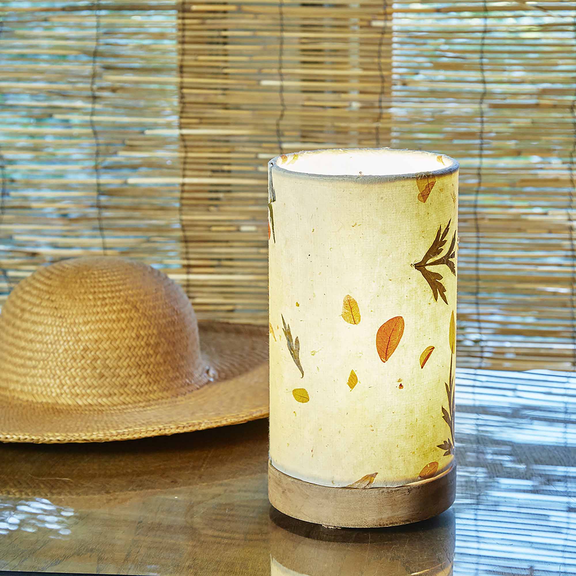 In a quiet bedside corner, crafted by the artisans of Eangee Home Design, the Mini Paper Cylinder Table Lamp Fern in natural stands gracefully, featuring reclaimed mango wood base, recycled paper shade, sustainably sourced, recycled materials, adding warmth and character to the space.