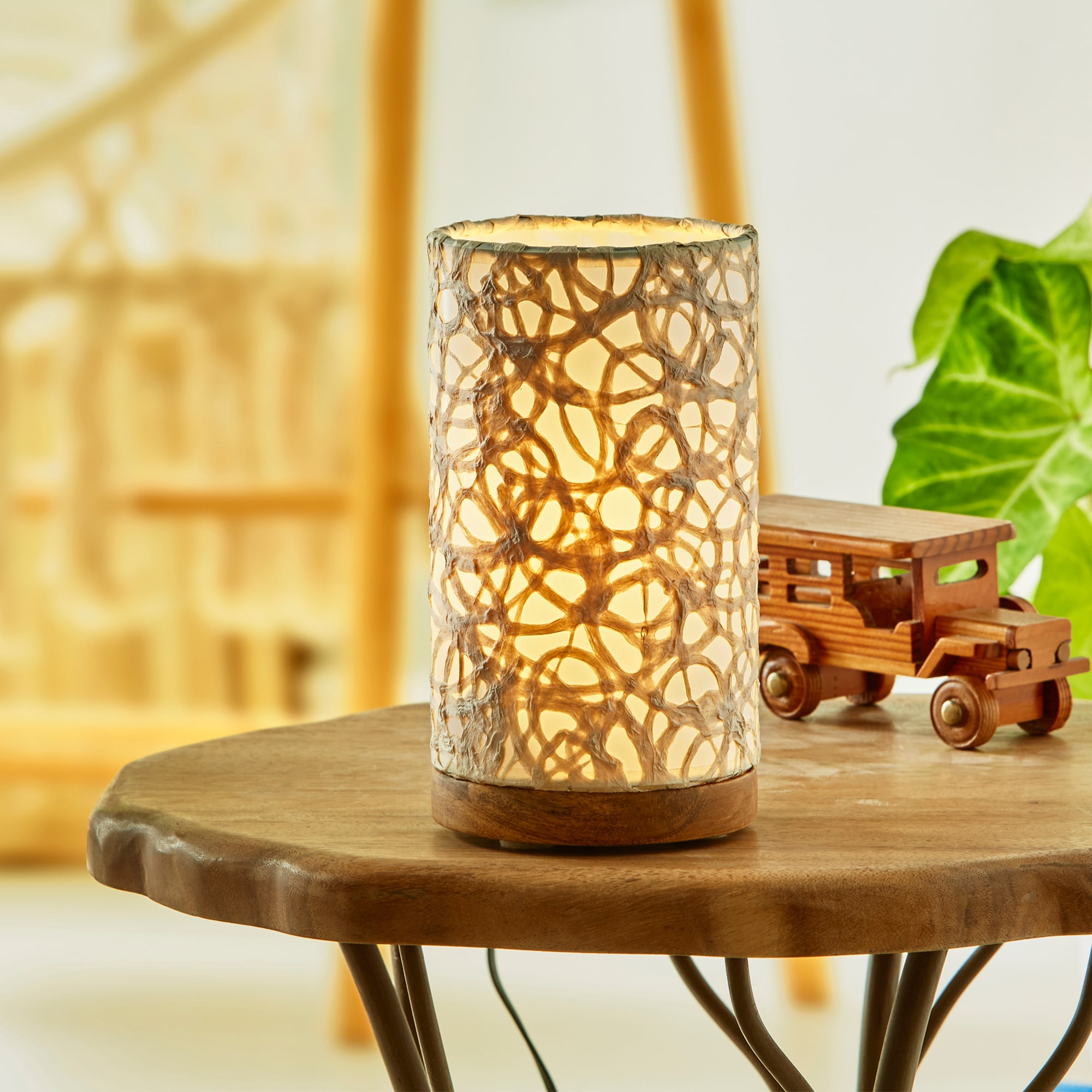 In a softly lit reading nook, crafted by the artisans of Eangee Home Design, the Mini Paper Cylinder Table Lamp Swirl in natural stands gracefully, featuring reclaimed mango wood base, recycled paper shade, sustainably sourced, recycled materials, adding warmth and character to the space.
