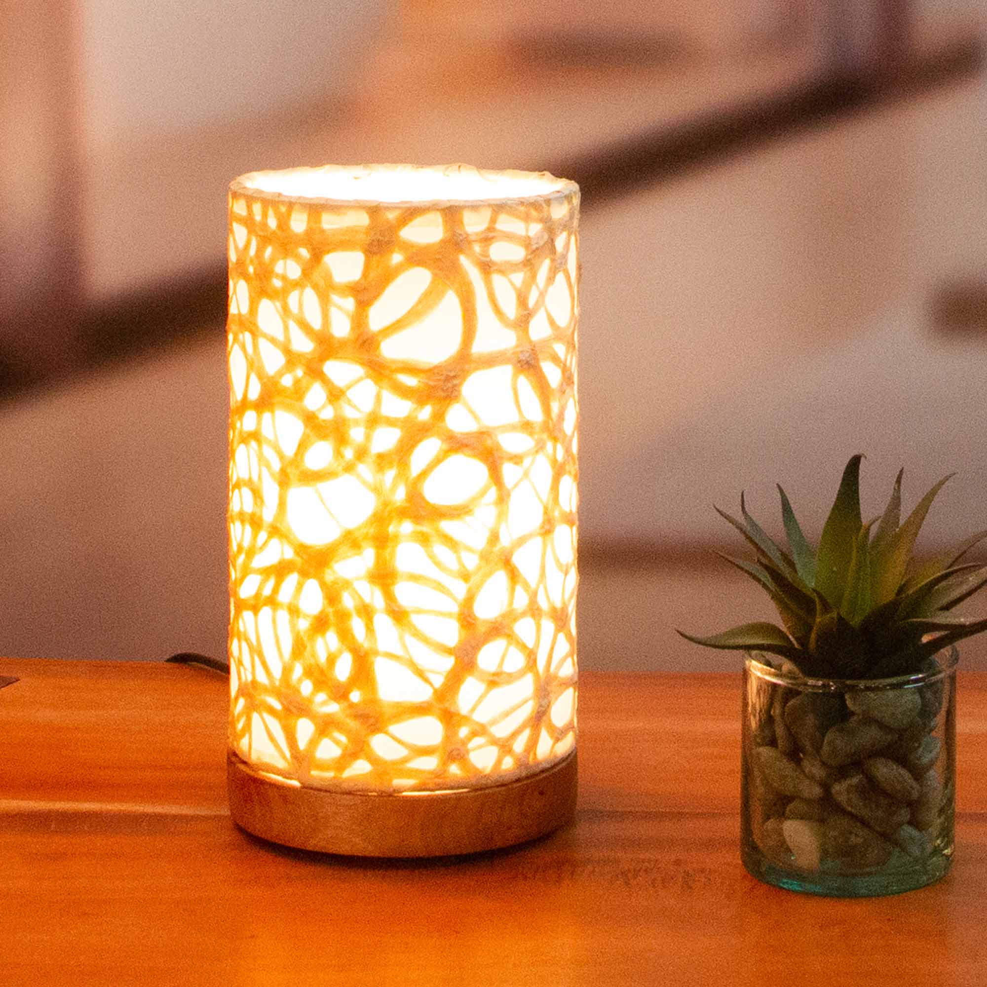 In a softly lit reading nook, crafted by the artisans of Eangee Home Design, the Mini Paper Cylinder Table Lamp Swirl in natural stands gracefully, featuring reclaimed mango wood base, recycled paper shade, sustainably sourced, recycled materials, adding warmth and character to the space.