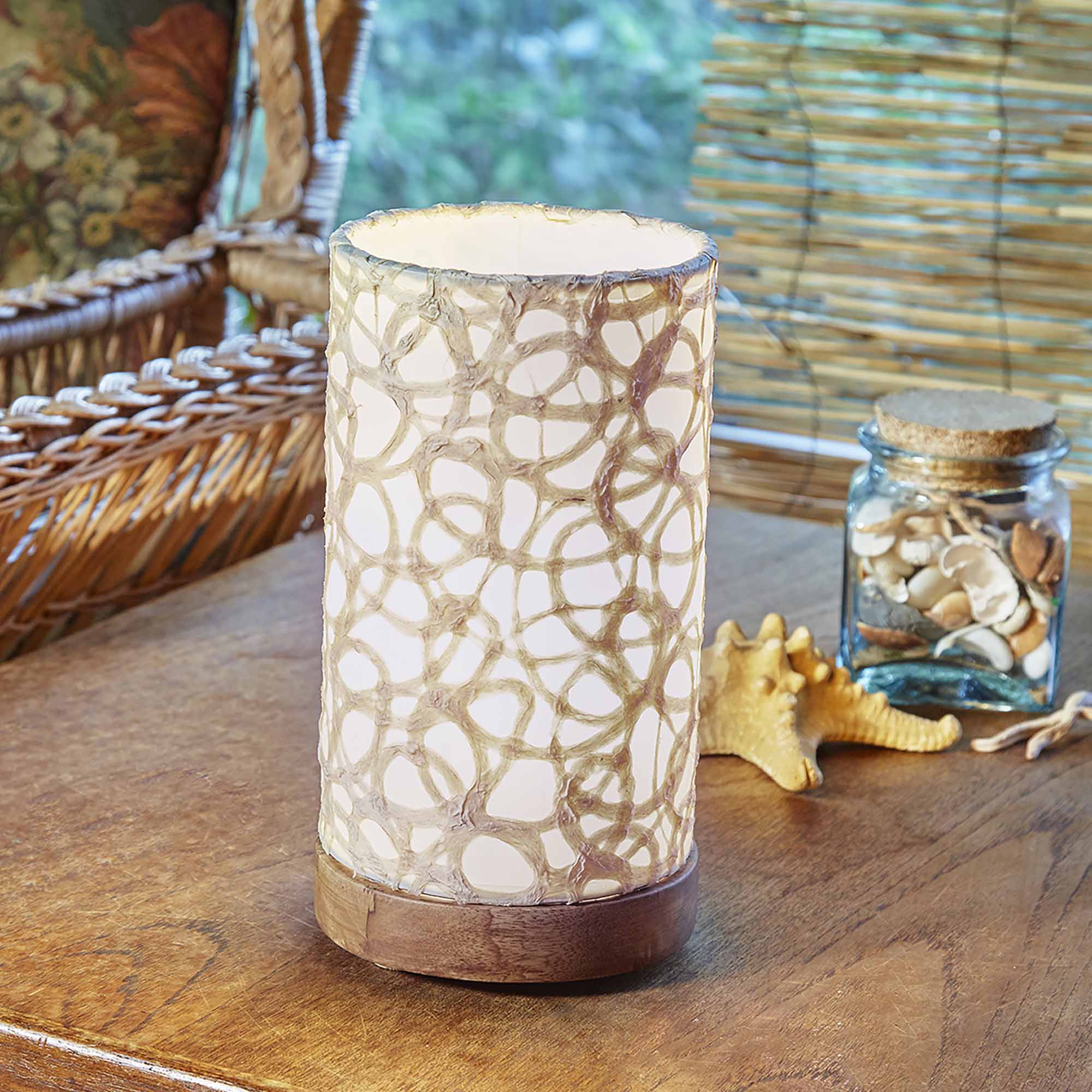 In a coastal inspired living room, crafted by the artisans of Eangee Home Design, the Mini Paper Cylinder Table Lamp Swirl in natural stands gracefully, featuring reclaimed mango wood base, recycled paper shade, sustainably sourced, recycled materials, adding warmth and character to the space.