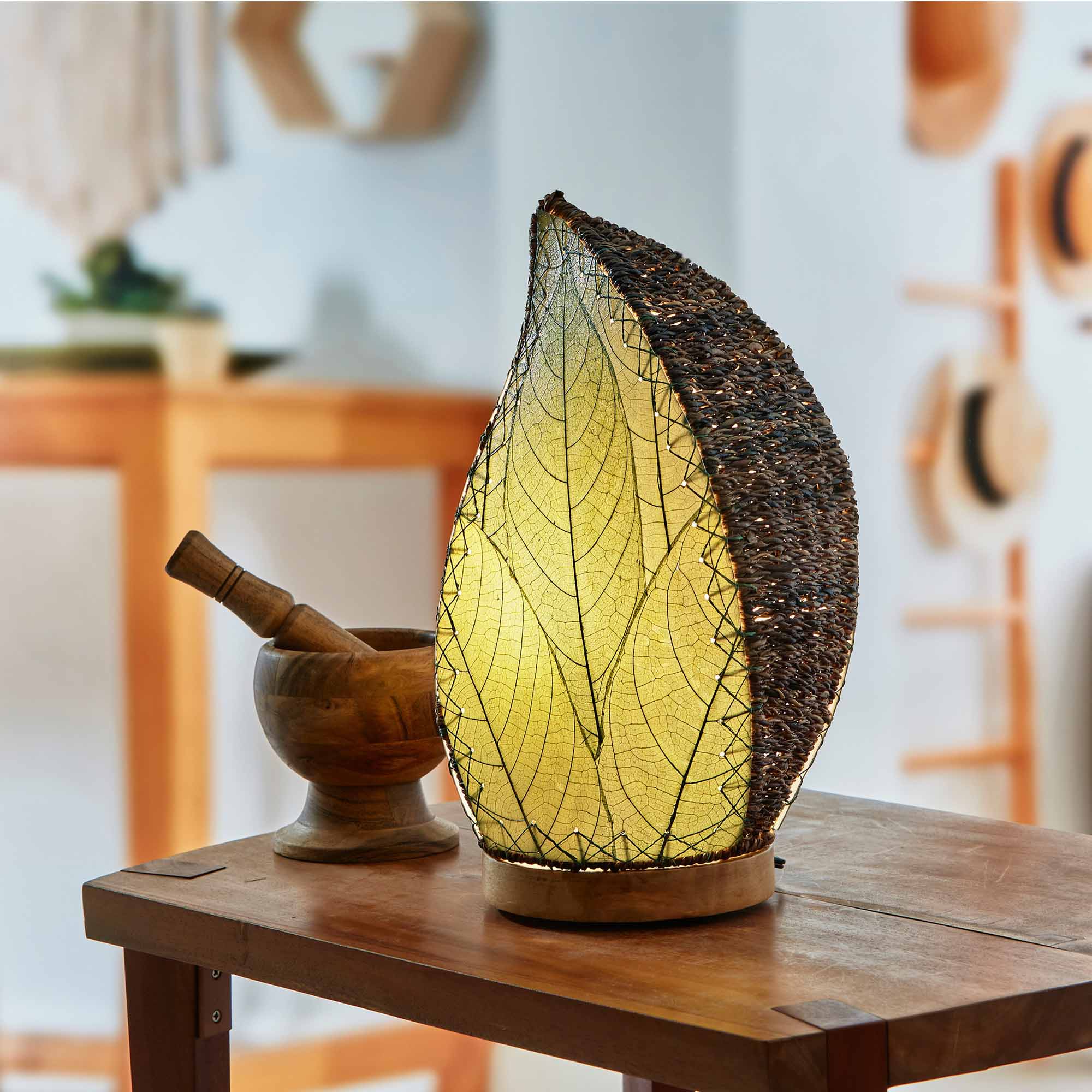 In a sun drenched entryway, crafted by the artisans of Eangee Home Design, the Leaflet Table Lamp in green stands gracefully, featuring fossilized cocoa leaf shade, abaca rope accents, reclaimed mango wood base, wrought iron frame, sustainably sourced, adding warmth and character to the space.