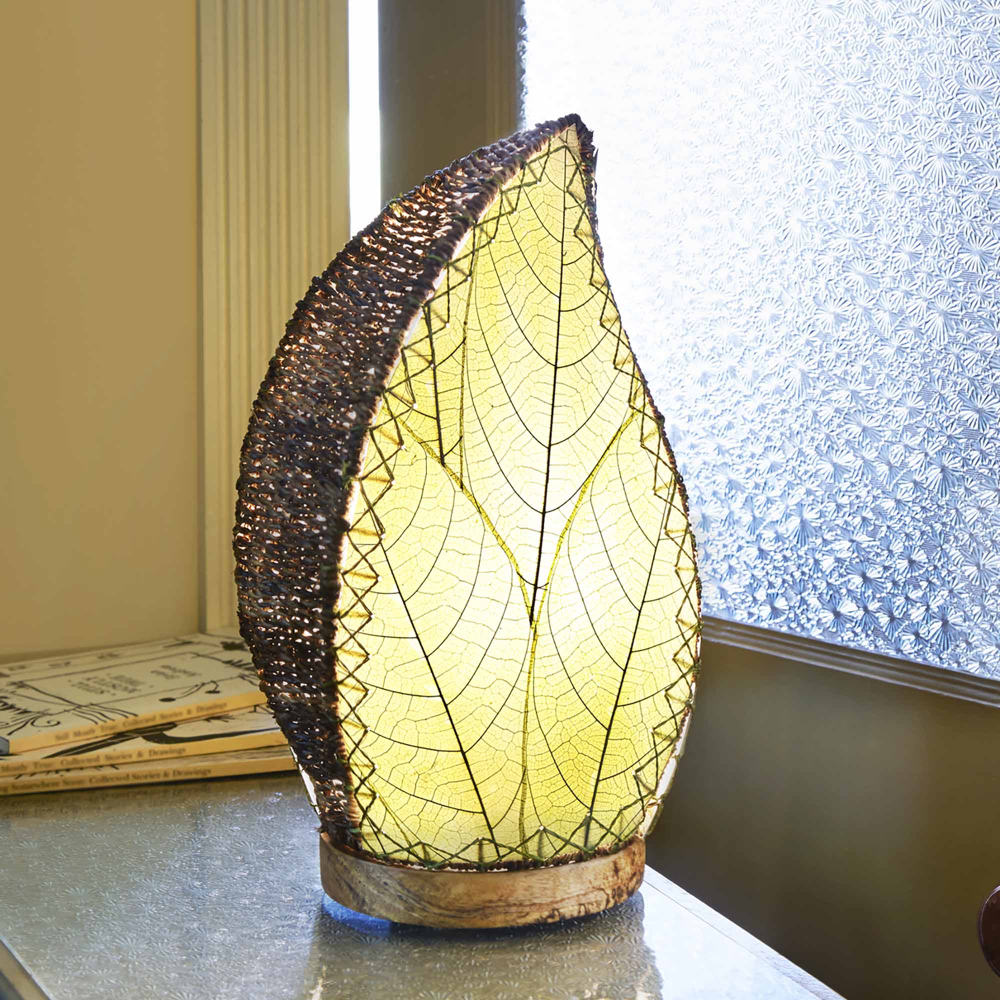 In a minimalist loft with natural textures, crafted by the artisans of Eangee Home Design, the Leaflet Table Lamp in green stands gracefully, featuring fossilized cocoa leaf shade, abaca rope accents, reclaimed mango wood base, wrought iron frame, sustainably sourced, adding warmth and character to the space.