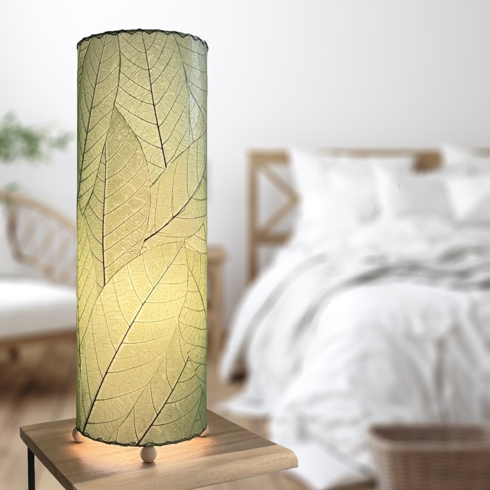 In a softly lit reading nook, crafted by the artisans of Eangee Home Design, the 24 Inch Cocoa Leaf Cylinder Table Lamp in green stands gracefully, featuring fossilized cocoa leaf shade, fair trade, eco friendly, sustainably sourced, handcrafted, adding warmth and character to the space.