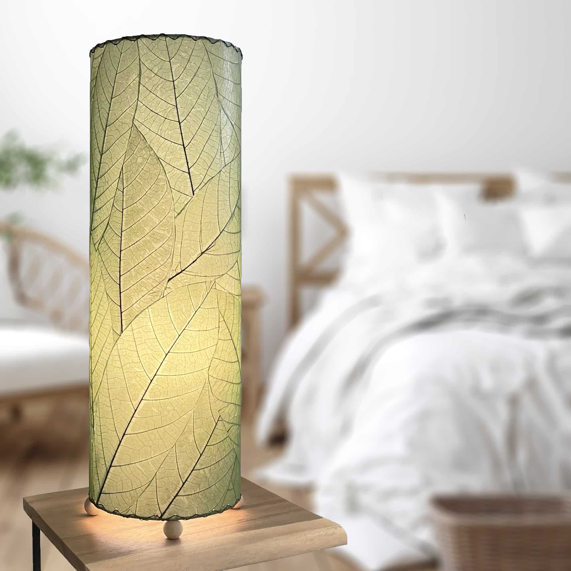 In a softly lit reading nook, crafted by the artisans of Eangee Home Design, the 24 Inch Cocoa Leaf Cylinder Table Lamp in green stands gracefully, featuring fossilized cocoa leaf shade, fair trade, eco friendly, sustainably sourced, handcrafted, adding warmth and character to the space.