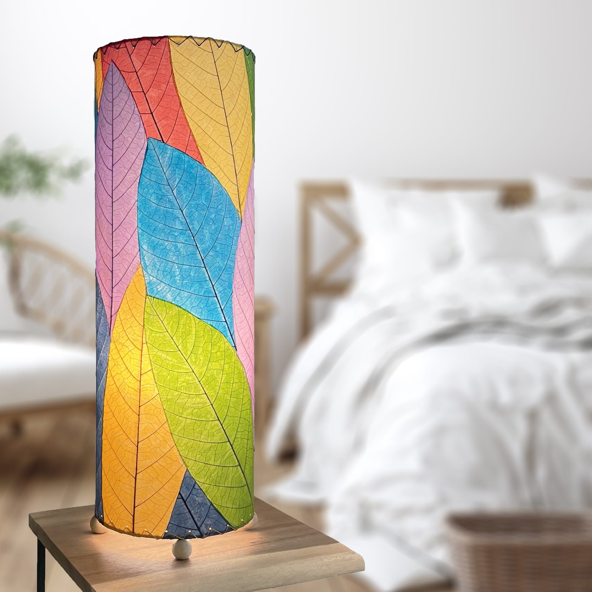 In a softly lit reading nook, crafted by the artisans of Eangee Home Design, the 24 Inch Cocoa Leaf Cylinder Table Lamp in multi color stands gracefully, featuring fossilized cocoa leaf shade, fair trade, eco friendly, sustainably sourced, handcrafted, adding warmth and character to the space.