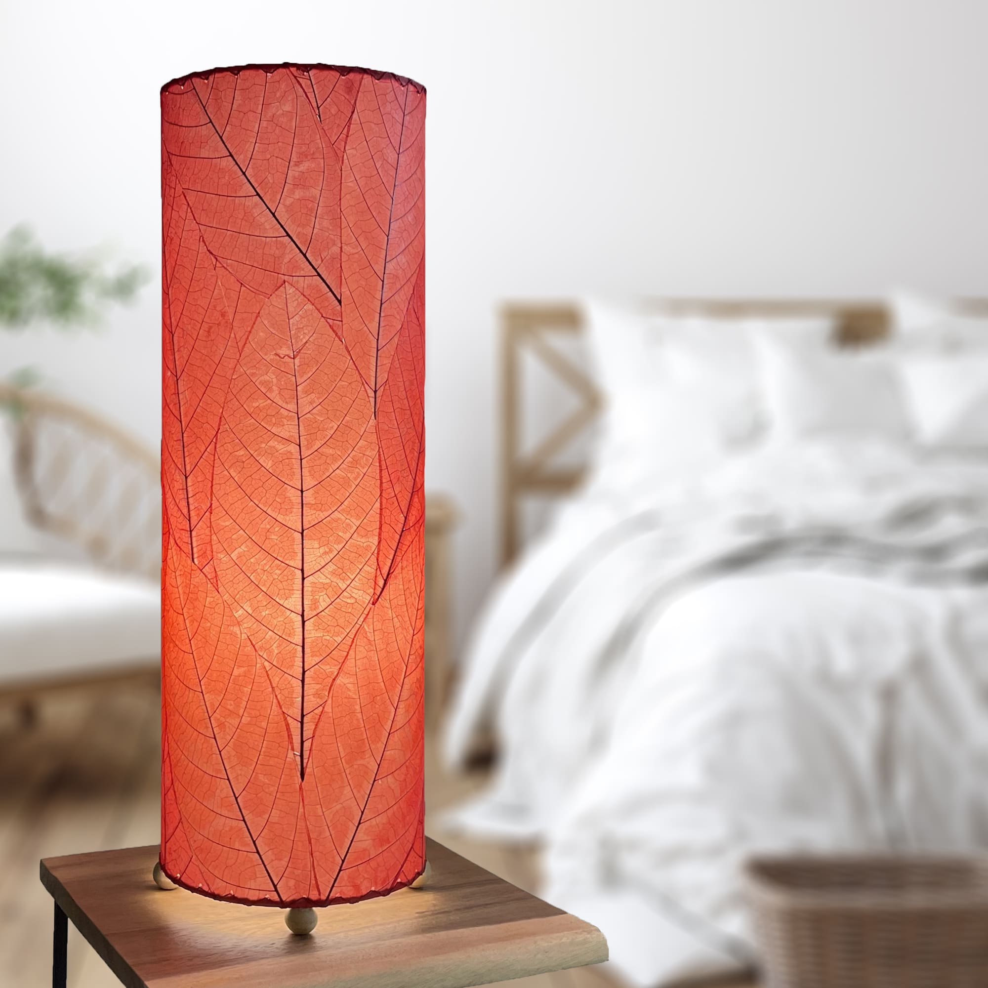 In a softly lit reading nook, crafted by the artisans of Eangee Home Design, the 24 Inch Cocoa Leaf Cylinder Table Lamp in red stands gracefully, featuring fossilized cocoa leaf shade, fair trade, eco friendly, sustainably sourced, handcrafted, adding warmth and character to the space.