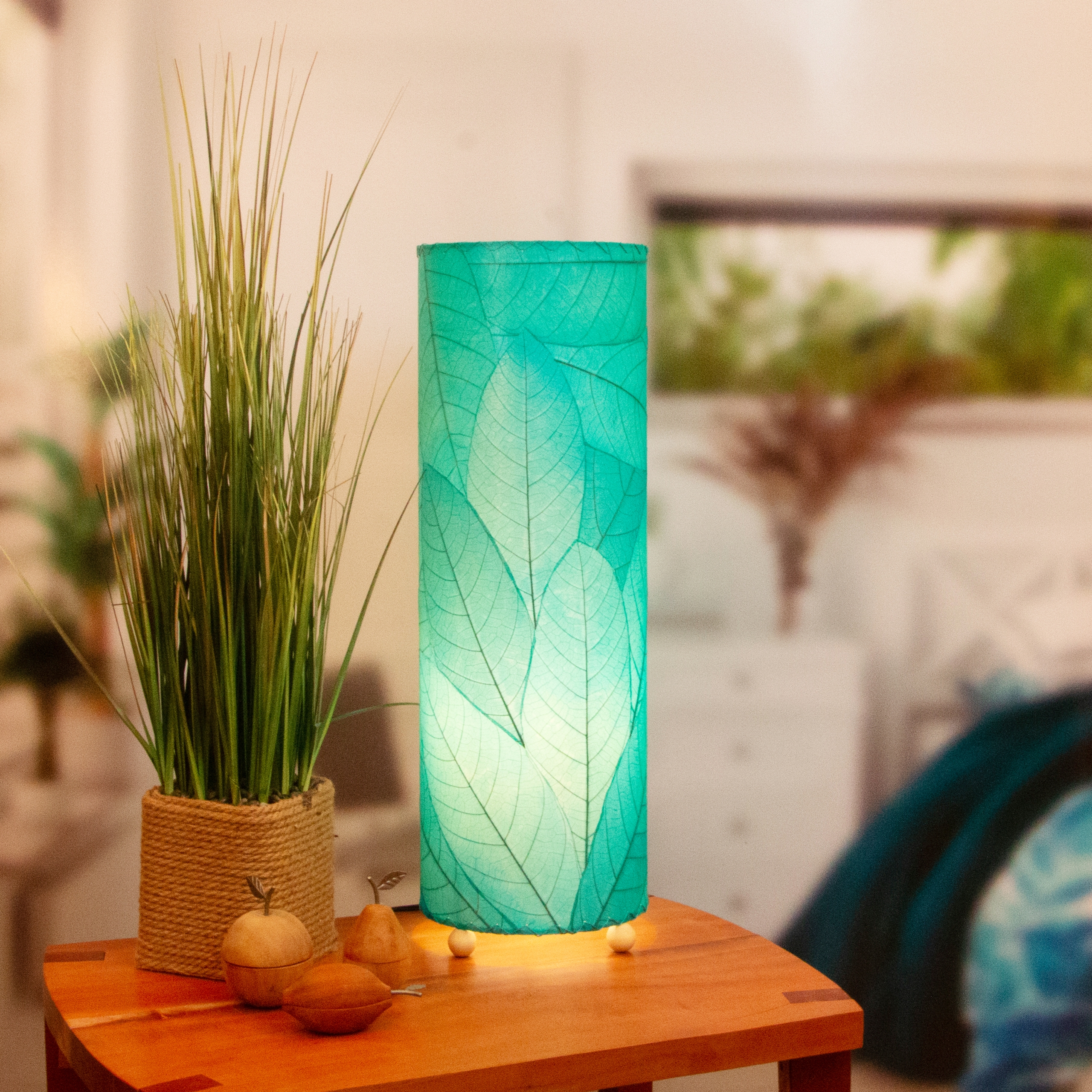 In a quiet bedside corner, crafted by the artisans of Eangee Home Design, the 24 Inch Cocoa Leaf Cylinder Table Lamp in sea blue stands gracefully, featuring fossilized cocoa leaf shade, fair trade, eco friendly, sustainably sourced, handcrafted, adding warmth and character to the space.