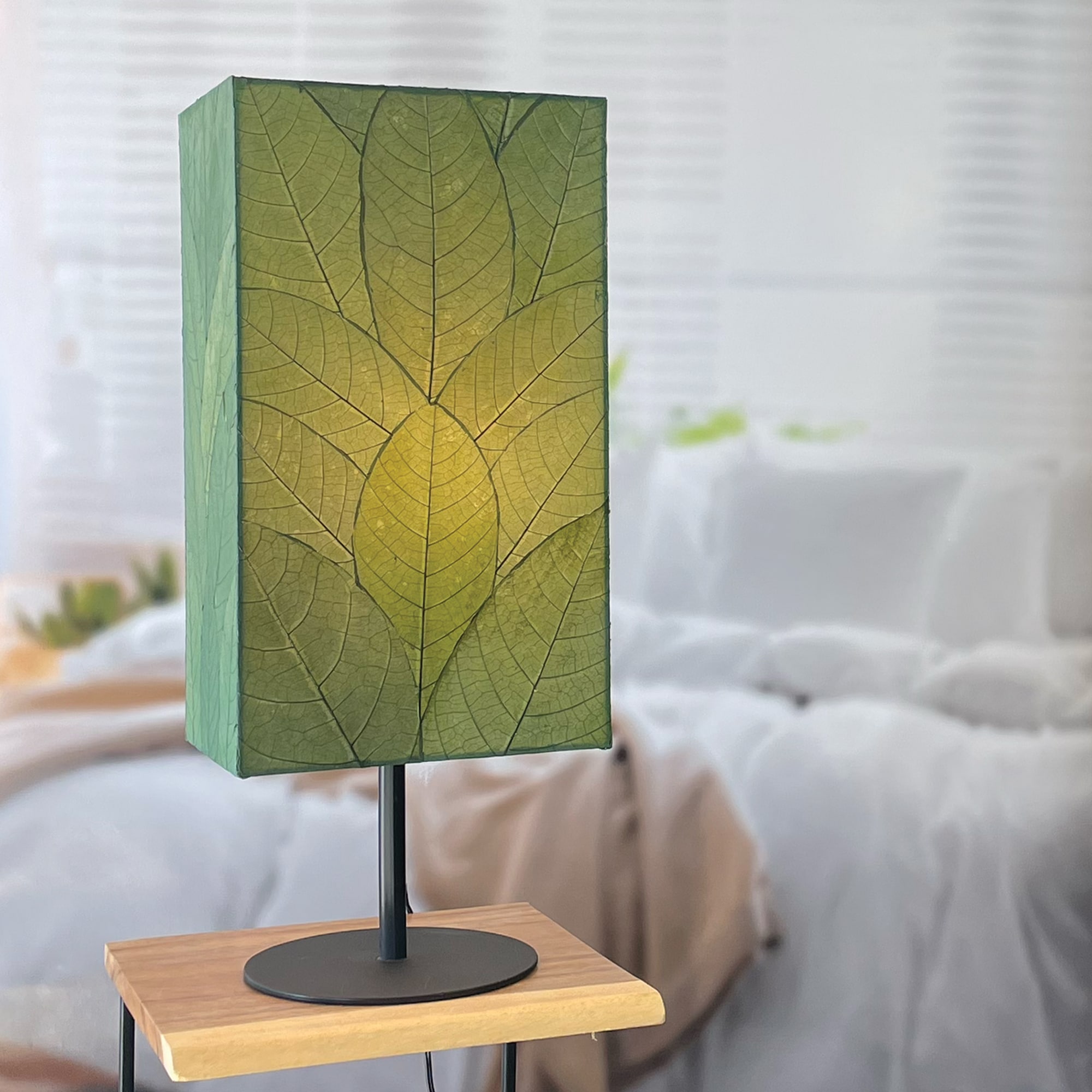 In a softly lit reading nook, crafted by the artisans of Eangee Home Design, the Sequoia Table Lamp in green stands gracefully, featuring fossilized cocoa leaf shade, fair trade, sustainably sourced, adding warmth and character to the space.
