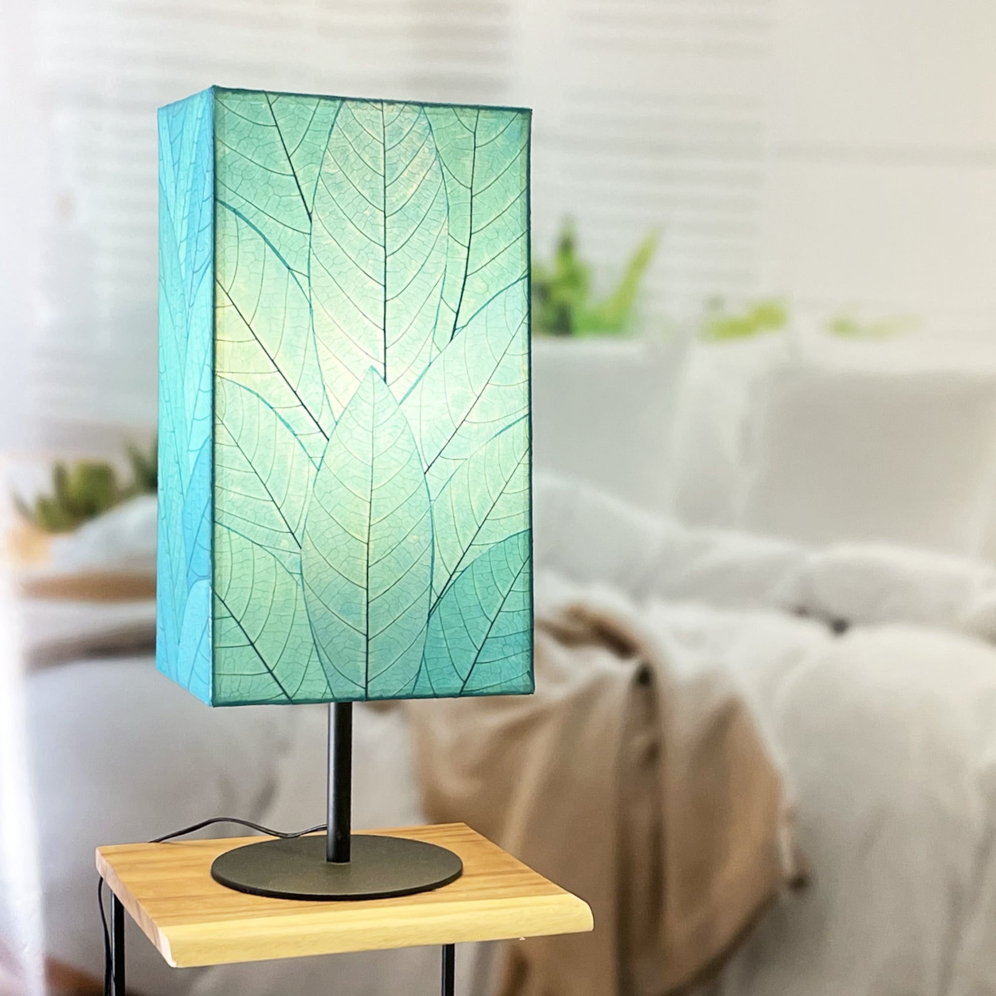 In a sun drenched entryway, crafted by the artisans of Eangee Home Design, the Sequoia Table Lamp in sea blue stands gracefully, featuring fossilized cocoa leaf shade, fair trade, sustainably sourced, adding warmth and character to the space.