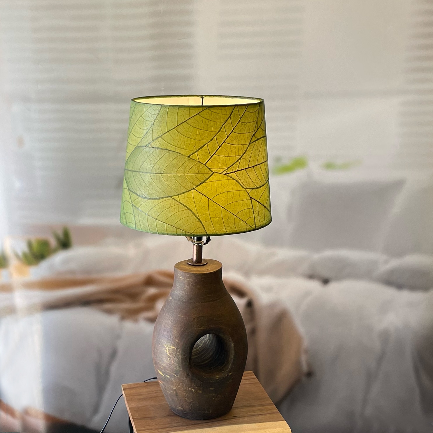 In a softly lit reading nook, crafted by the artisans of Eangee Home Design, the 12 Inch Tapered Drum Shade in green stands gracefully, featuring fossilized cocoa leaf shade, fair trade, adding warmth and character to the space.