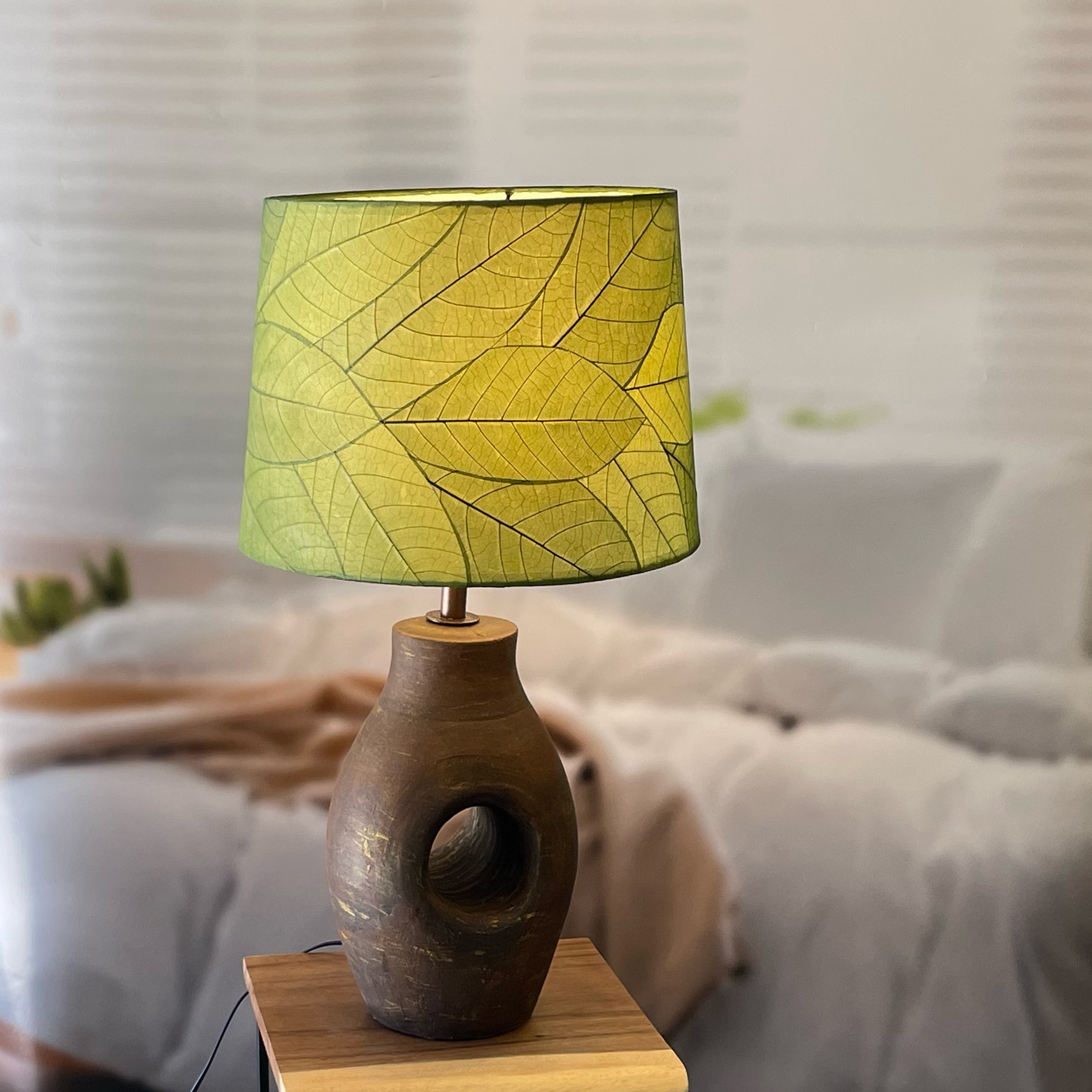 In a modern interior with earth tones, crafted by the artisans of Eangee Home Design, the 14 Inch Tapered Drum Shade in green stands gracefully, featuring fossilized cocoa leaf shade, wrought iron frame, fair trade, eco friendly, sustainably sourced, handcrafted, adding warmth and character to the space.