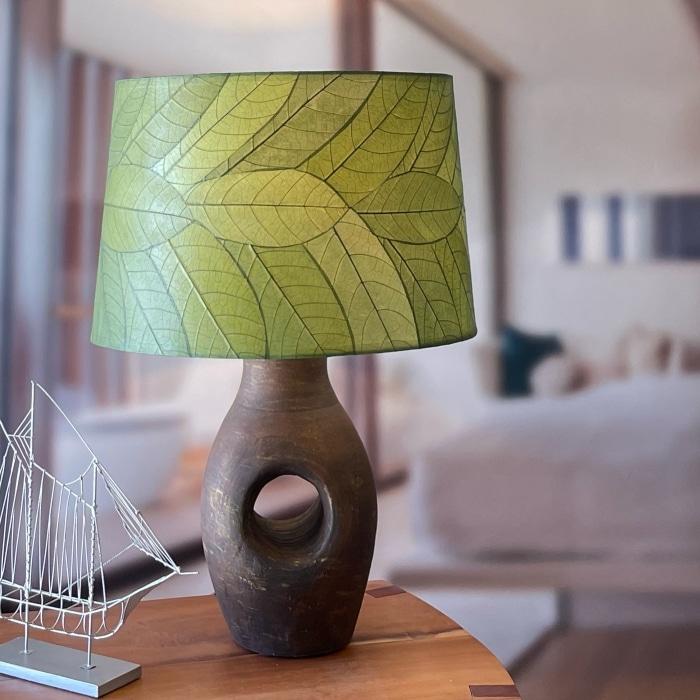 In a sun drenched entryway, crafted by the artisans of Eangee Home Design, the 18 Inch Tapered Drum Shade in green stands gracefully, featuring fossilized cocoa leaf shade, wrought iron frame, eco friendly, sustainably sourced, adding warmth and character to the space.
