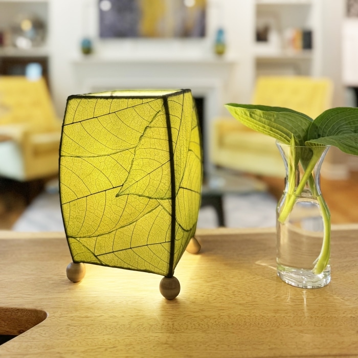 In a softly lit reading nook, crafted by the artisans of Eangee Home Design, the Mini Square Cocoa Leaf Table Lamp in green stands gracefully, featuring fossilized cocoa leaf shade, wrought iron frame, fair trade, eco friendly, sustainably sourced, handcrafted, adding warmth and character to the space.