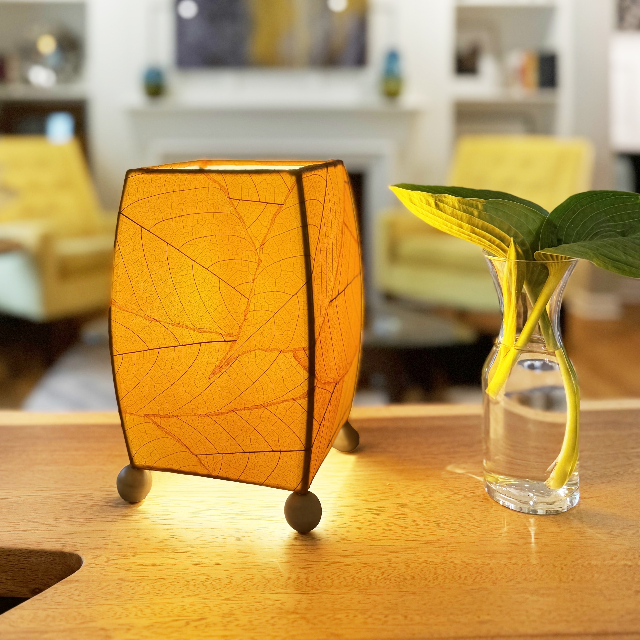 In a sun drenched entryway, crafted by the artisans of Eangee Home Design, the Mini Square Cocoa Leaf Table Lamp in orange stands gracefully, featuring fossilized cocoa leaf shade, wrought iron frame, fair trade, eco friendly, sustainably sourced, handcrafted, adding warmth and character to the space.