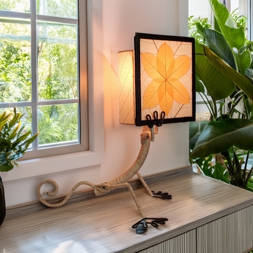 In a sun drenched entryway, crafted by the artisans of Eangee Home Design, the Enlightened Gecko Table Lamp in orange stands gracefully, featuring fossilized cocoa leaf shade, guyabano leaf shade, abaca rope accents, wrought iron frame, handcrafted, adding warmth and character to the space.