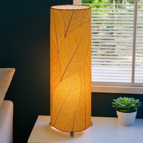 In a modern interior with earth tones, crafted by the artisans of Eangee Home Design, the 24 Inch Cocoa Leaf Cylinder Table Lamp in orange stands gracefully, featuring fossilized cocoa leaf shade, fair trade, eco friendly, sustainably sourced, handcrafted, adding warmth and character to the space.