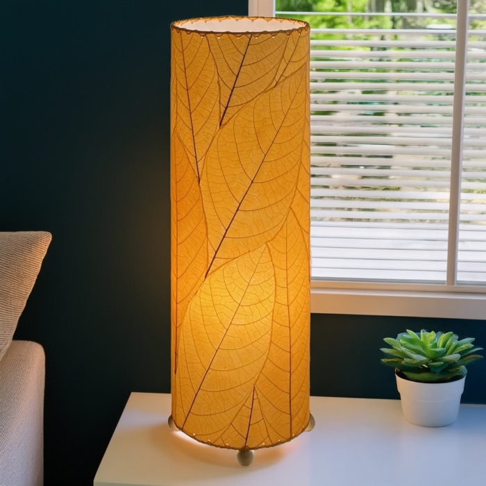 In a modern interior with earth tones, crafted by the artisans of Eangee Home Design, the 24 Inch Cocoa Leaf Cylinder Table Lamp in orange stands gracefully, featuring fossilized cocoa leaf shade, fair trade, eco friendly, sustainably sourced, handcrafted, adding warmth and character to the space.