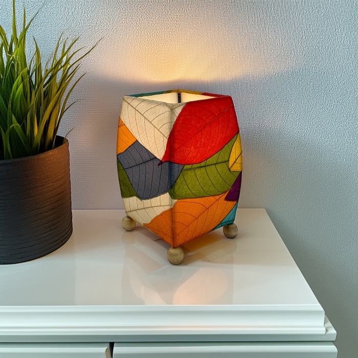 In a quiet bedside corner, crafted by the artisans of Eangee Home Design, the Mini Square Cocoa Leaf Table Lamp in multi color stands gracefully, featuring fossilized cocoa leaf shade, wrought iron frame, fair trade, eco friendly, sustainably sourced, handcrafted, adding warmth and character to the space.