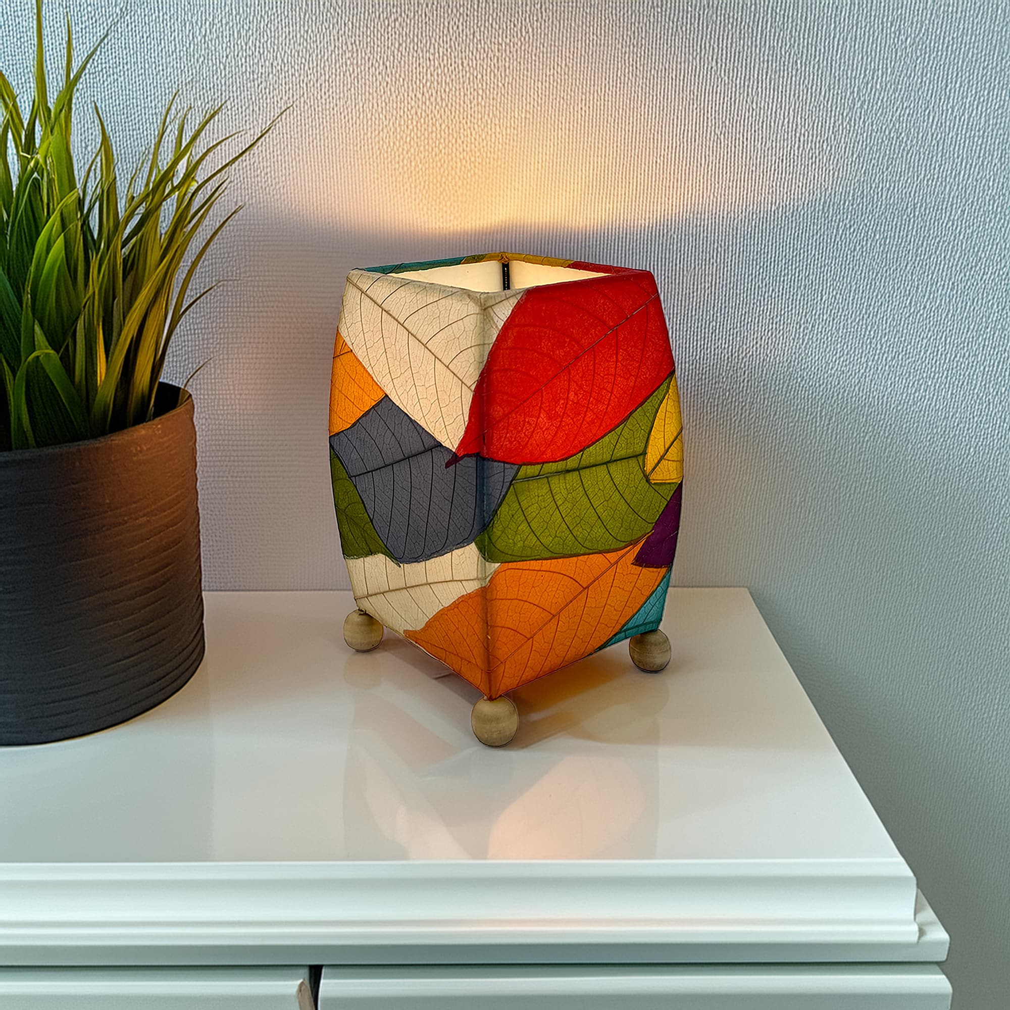 In a quiet bedside corner, crafted by the artisans of Eangee Home Design, the Mini Square Cocoa Leaf Table Lamp in multi color stands gracefully, featuring fossilized cocoa leaf shade, wrought iron frame, fair trade, eco friendly, sustainably sourced, handcrafted, adding warmth and character to the space.