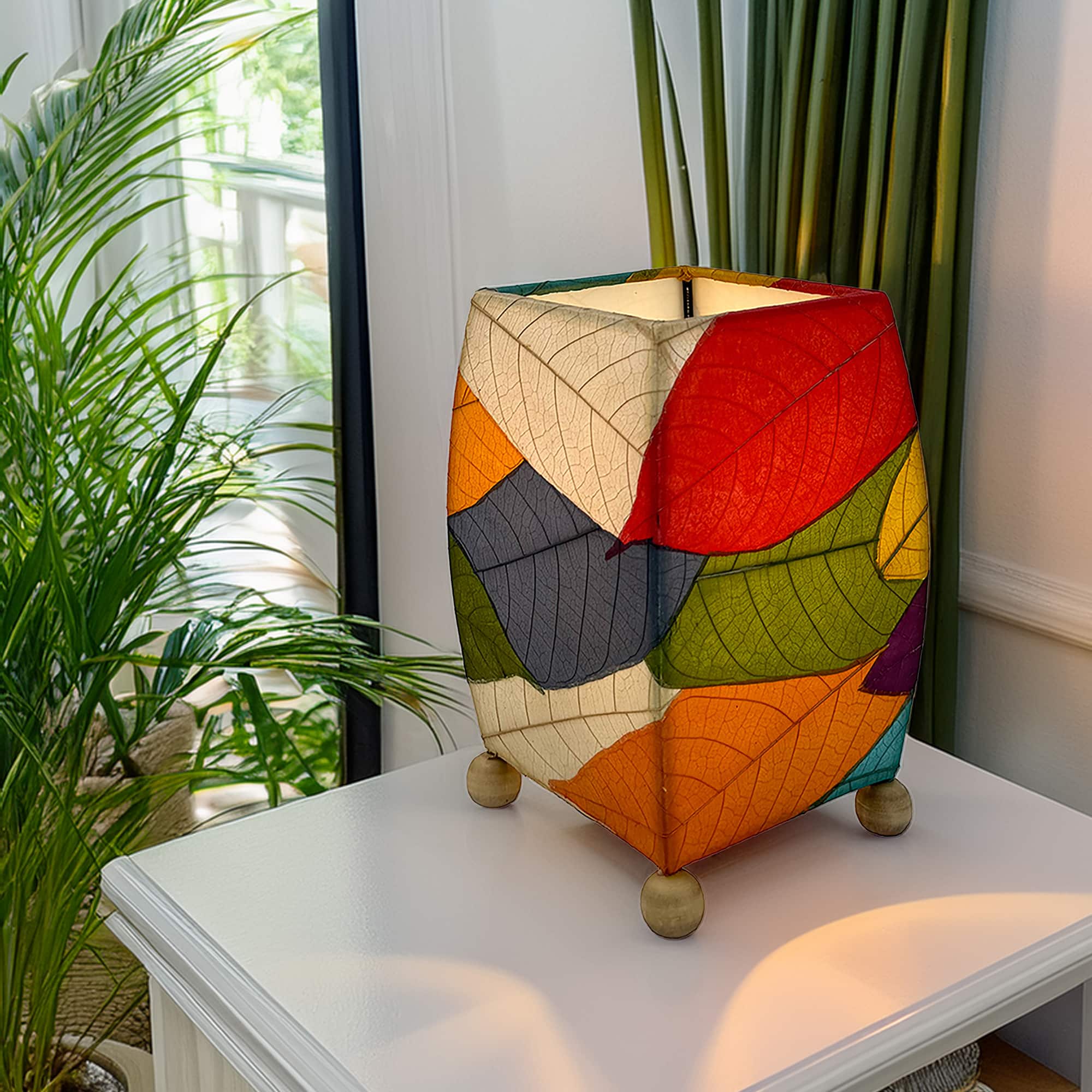 In a quiet bedside corner, crafted by the artisans of Eangee Home Design, the Mini Square Cocoa Leaf Table Lamp in multi color stands gracefully, featuring fossilized cocoa leaf shade, wrought iron frame, fair trade, eco friendly, sustainably sourced, handcrafted, adding warmth and character to the space.