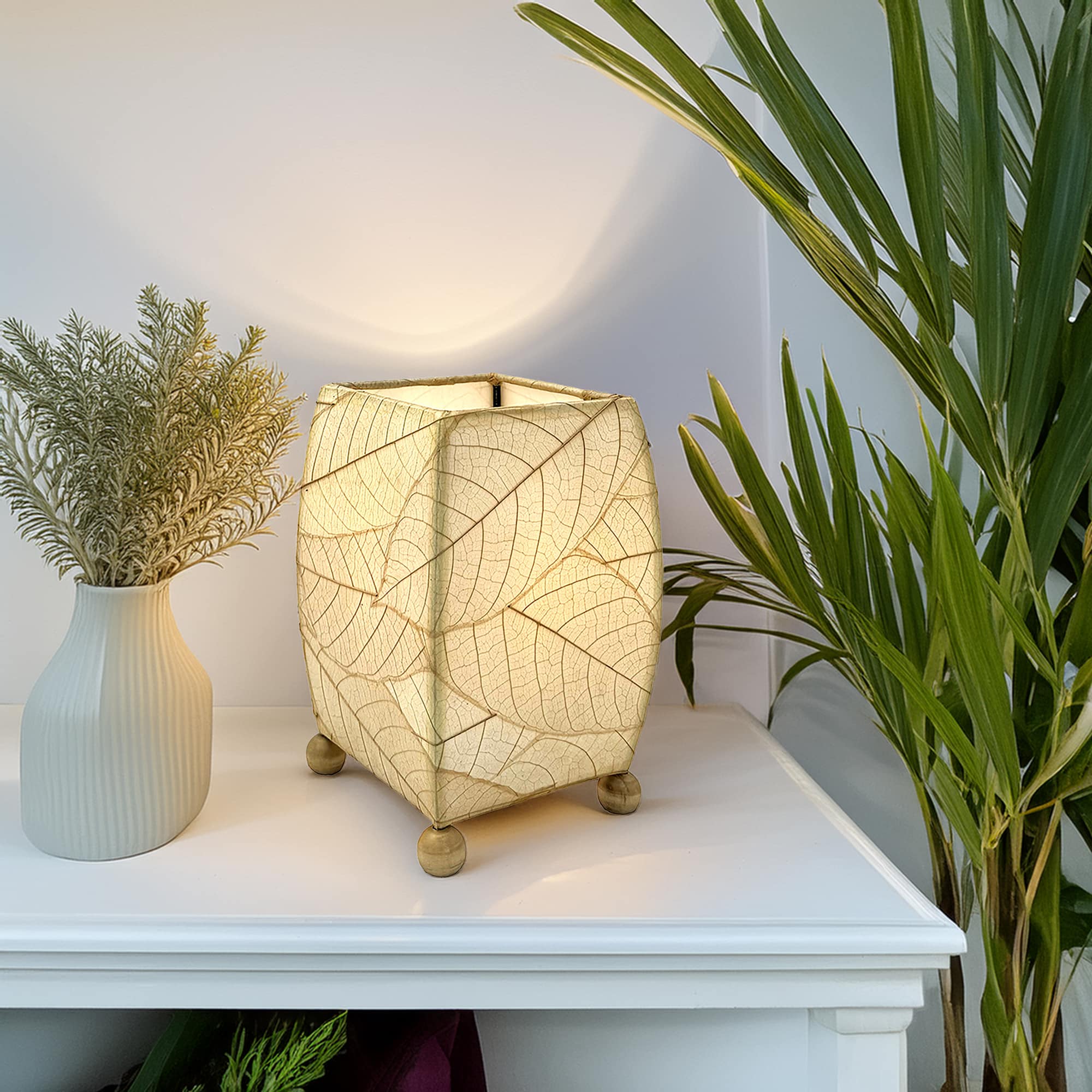 In a sun drenched entryway, crafted by the artisans of Eangee Home Design, the Mini Square Cocoa Leaf Table Lamp in natural stands gracefully, featuring fossilized cocoa leaf shade, wrought iron frame, fair trade, eco friendly, sustainably sourced, handcrafted, adding warmth and character to the space.