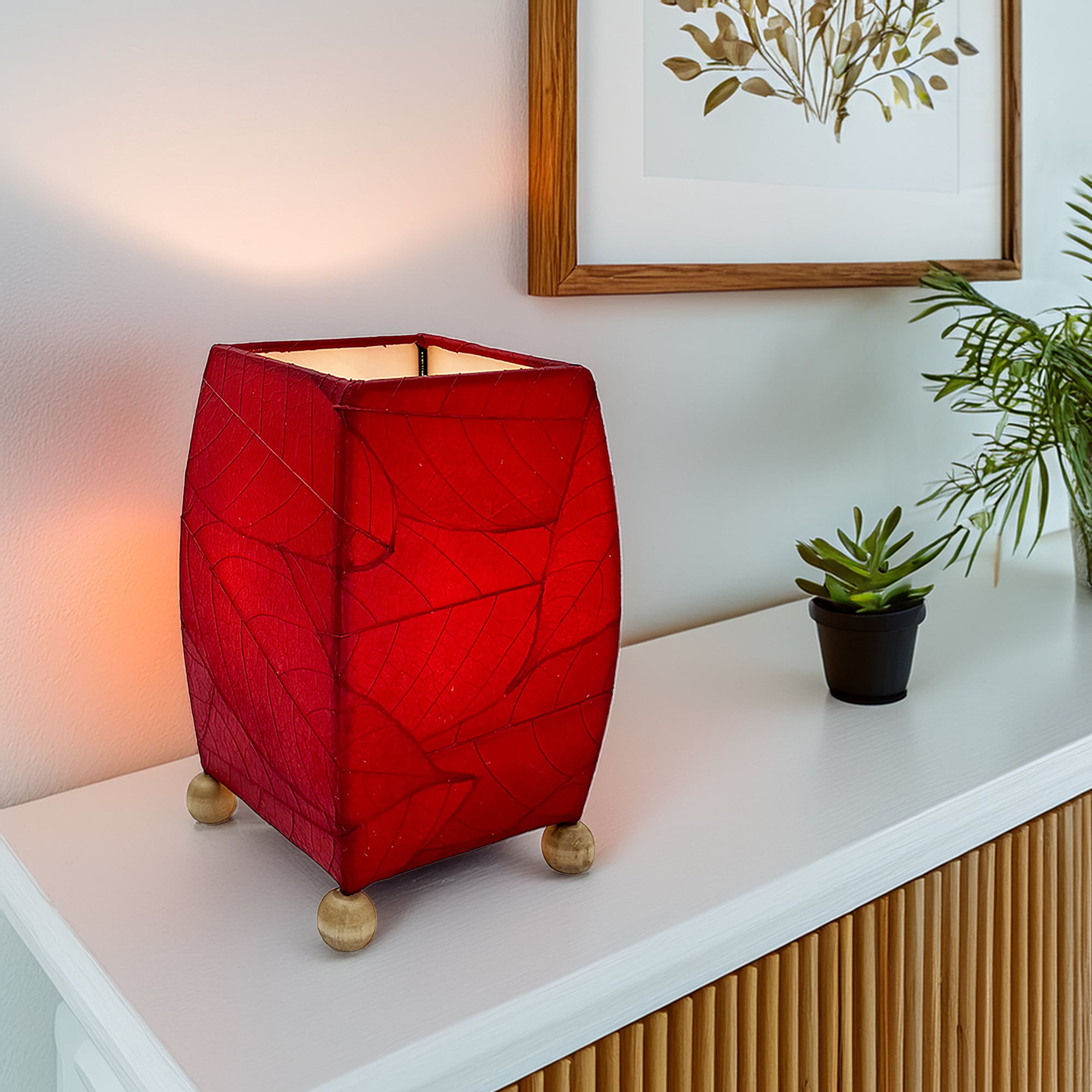 In a softly lit reading nook, crafted by the artisans of Eangee Home Design, the Mini Square Cocoa Leaf Table Lamp in red stands gracefully, featuring fossilized cocoa leaf shade, wrought iron frame, fair trade, eco friendly, sustainably sourced, handcrafted, adding warmth and character to the space.