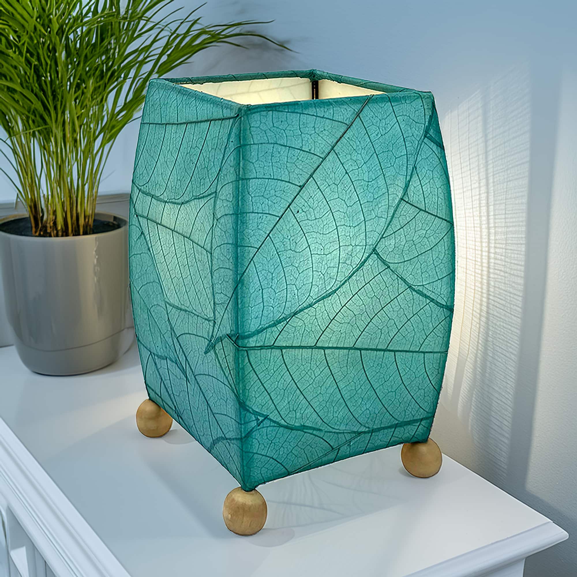 In a quiet bedside corner, crafted by the artisans of Eangee Home Design, the Mini Square Cocoa Leaf Table Lamp in sea blue stands gracefully, featuring fossilized cocoa leaf shade, wrought iron frame, fair trade, eco friendly, sustainably sourced, handcrafted, adding warmth and character to the space.