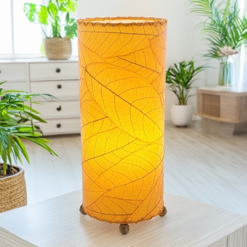 In a modern interior with earth tones, crafted by the artisans of Eangee Home Design, the Cocoa Leaf Cylinder Table Lamp in orange stands gracefully, featuring fossilized cocoa leaf shade, wrought iron frame, handcrafted, adding warmth and character to the space.