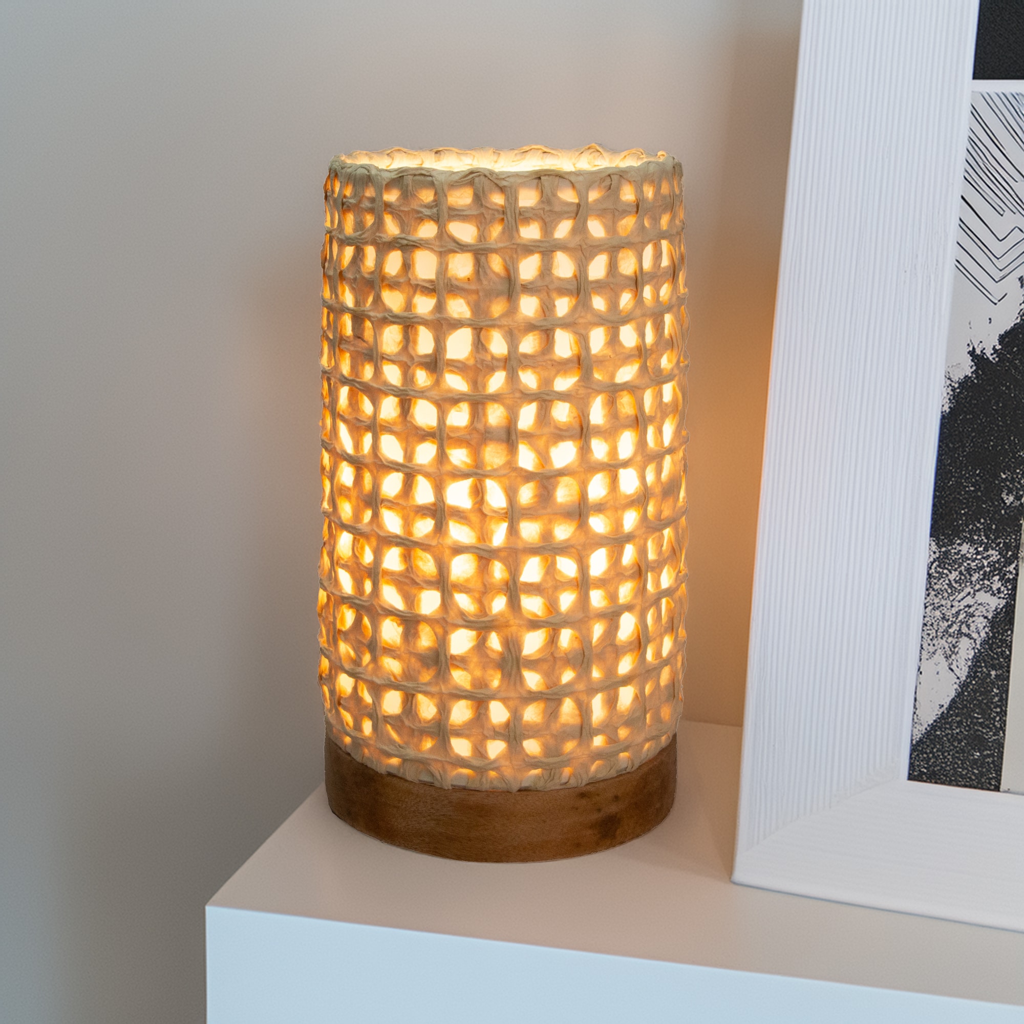 In a quiet bedside corner, crafted by the artisans of Eangee Home Design, the Mini Paper Cylinder Table Lamp Mesh in natural stands gracefully, featuring reclaimed mango wood base, recycled paper shade, sustainably sourced, recycled materials, adding warmth and character to the space.