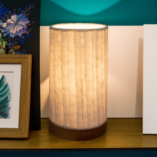In a softly lit reading nook, crafted by the artisans of Eangee Home Design, the Mini Paper Cylinder Table Lamp Lines in natural stands gracefully, featuring reclaimed mango wood base, recycled paper shade, sustainably sourced, recycled materials, adding warmth and character to the space.