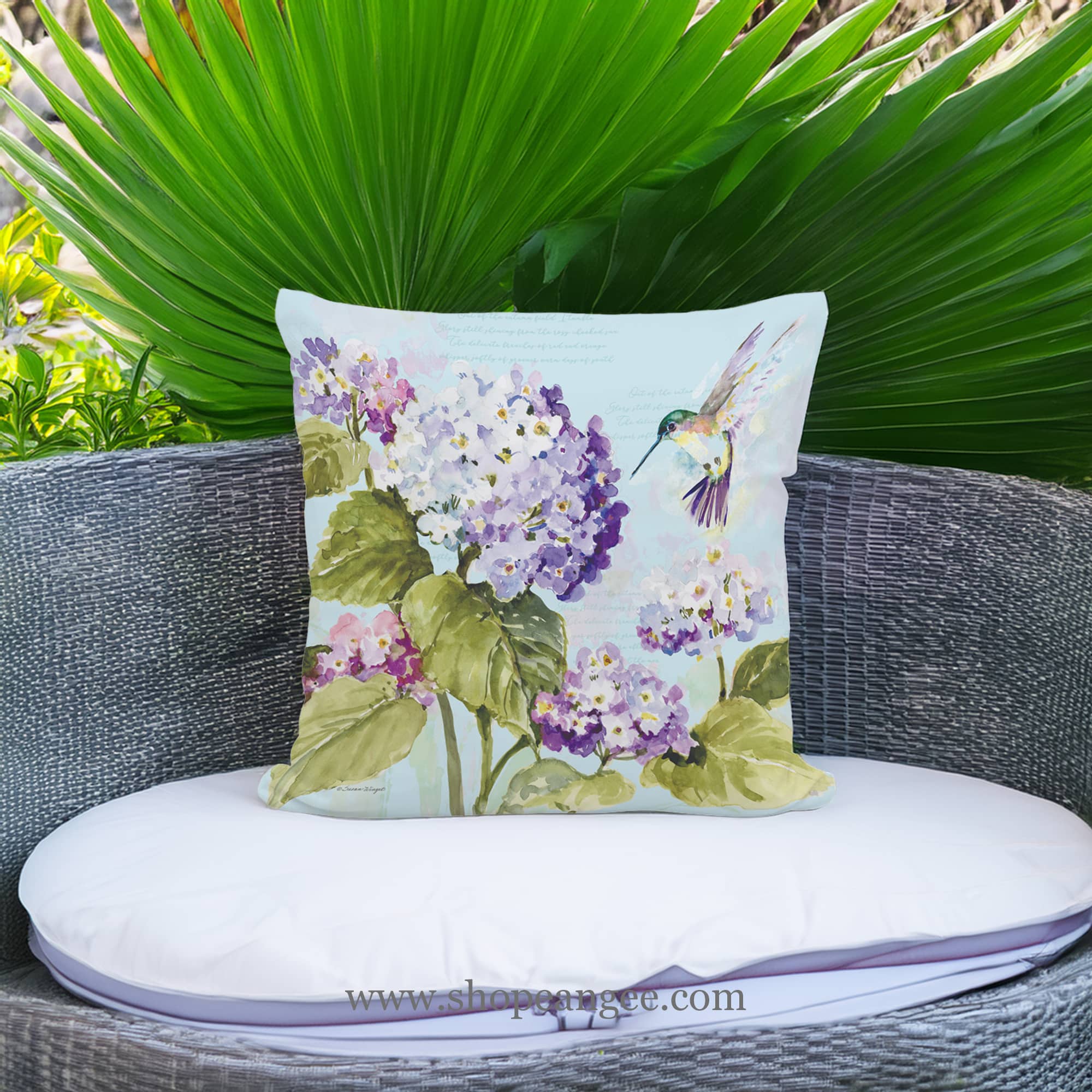 In an outdoor reading nook a multi colored outdoor pillow crafted by the artisans of Magnolia Casual called the Outdoor Pillow Hummingbird with Hydrangea 18x18 is displayed featuring handwoven textile adding comfort and style to the space featuring with a hummingbird motif.