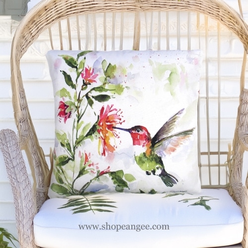 In a shaded porch swing a multi colored outdoor pillow crafted by the artisans of Magnolia Casual called the Outdoor Pillow Hummer w/Hanging Plant 2 18x18 is displayed featuring quality outdoor fabric adding comfort and style to the space.