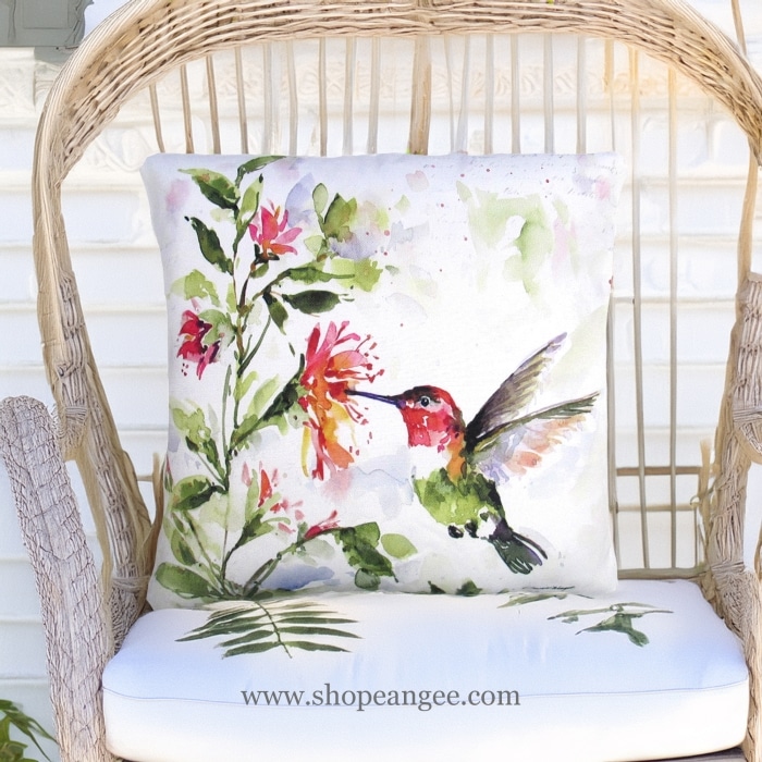 In a shaded porch swing a multi colored outdoor pillow crafted by the artisans of Magnolia Casual called the Outdoor Pillow Hummer w/Hanging Plant 2 18x18 is displayed featuring quality outdoor fabric adding comfort and style to the space.