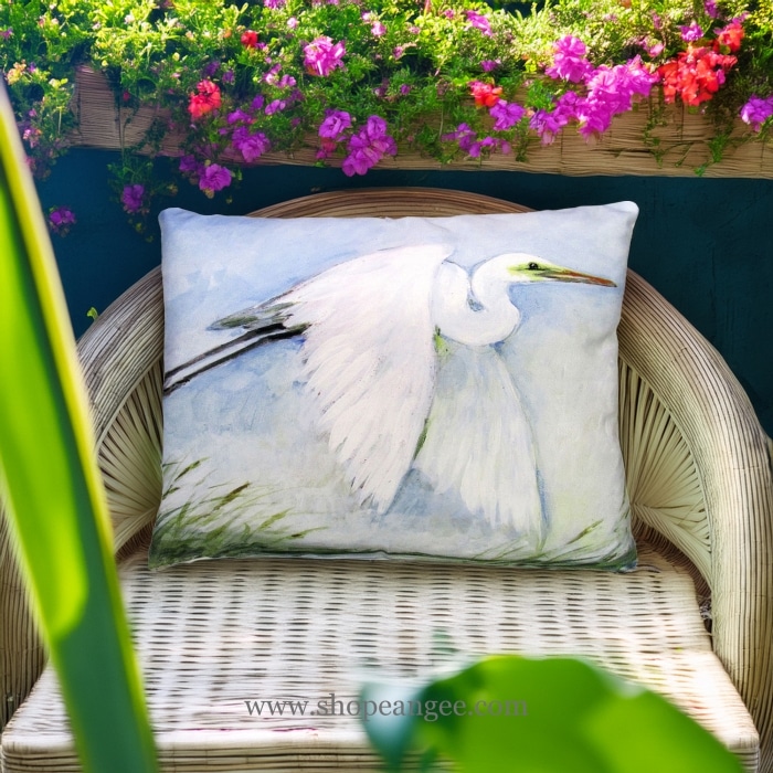 In a sunlit patio lounge a white outdoor pillow crafted by the artisans of Magnolia Casual called the Outdoor Pillow White Egret Flying 19x24 is displayed featuring quality outdoor fabric adding comfort and style to the space featuring shades of white.
