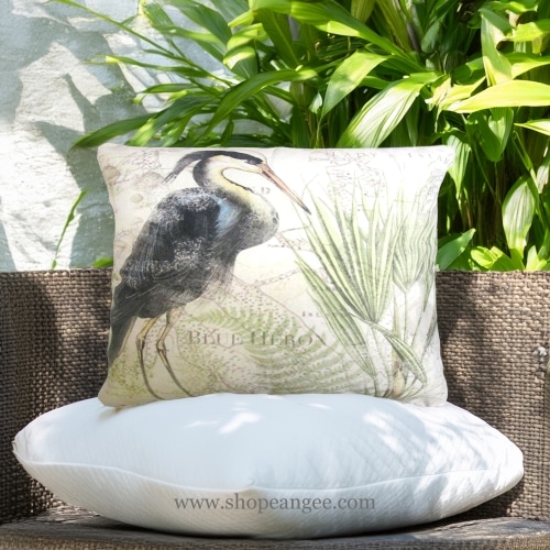 In an outdoor reading nook a multi colored outdoor pillow crafted by the artisans of Magnolia Casual called the Outdoor Pillow Great Heron 1 19x24 is displayed featuring quality outdoor fabric, natural materials adding comfort and style to the space.