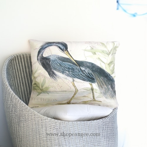 In an outdoor reading nook a multi colored outdoor pillow crafted by the artisans of Magnolia Casual called the Outdoor Pillow Great Heron 2 19x24 is displayed featuring quality outdoor fabric, natural materials adding comfort and style to the space.