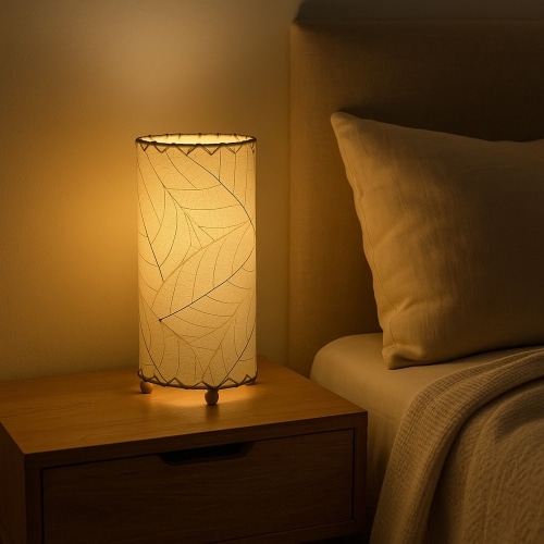 12 Inch Cocoa Leaf Cylinder Table Lamp Natural shade, handcrafted from real cocoa leaves . soft ambient light on a bedroom nightstand casting a warm glow next to a beige headboard.